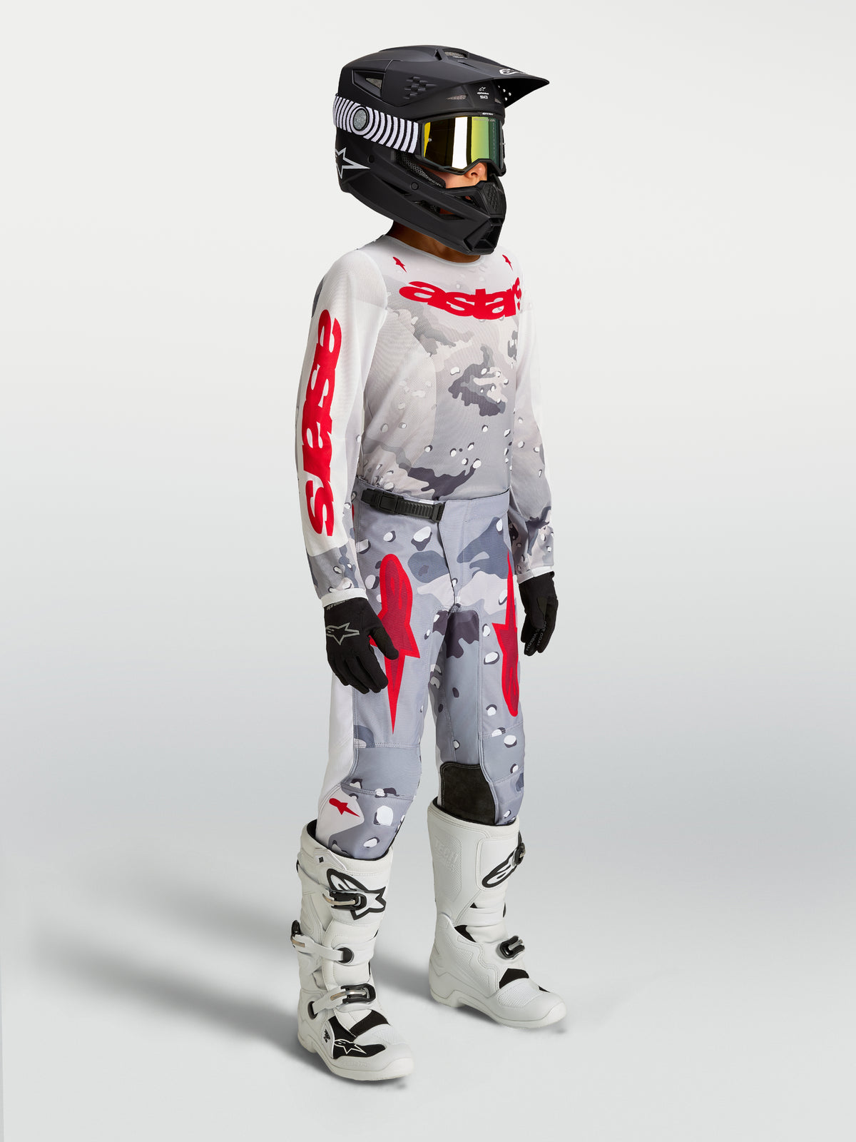 Alpinestars Jugend Fluid Lone Gear Set, Motocross-Trikot und -Hose in Grau-Camo und Rot, getragen mit weißen Stiefeln.