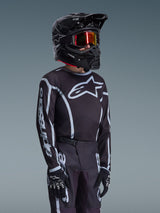 Alpinestars 2026 Youth Fluid Apex Jersey in Black and Gray, worn by a young rider with a matching black helmet and tinted goggles, featuring prominent Alpinestars branding on the chest and sleeves, part of a coordinated motocross gear set.