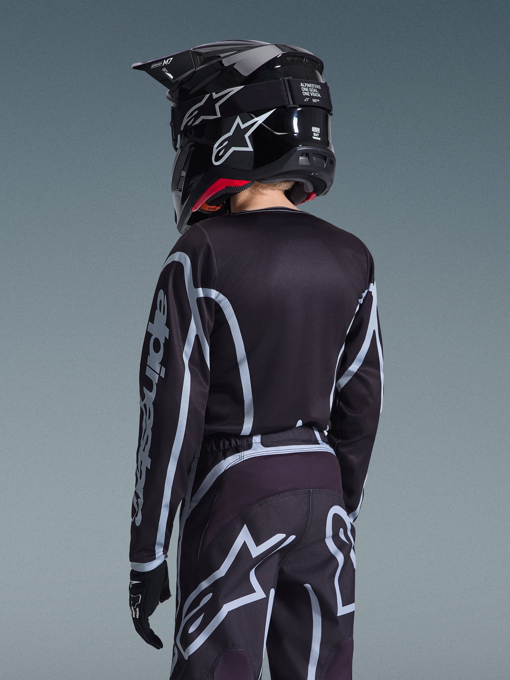 Rear view of a youth motocross rider wearing the Alpinestars 2026 Youth Fluid Apex Jersey and matching pants in Black and Gray, featuring a black helmet with white branding and black gloves, athletic off-road racing gear for young riders.