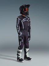 Youth motocross rider wearing the 2026 Youth Fluid Apex Jersey in Black Gray, matching black and gray pants with white graphics, black helmet with red-tinted goggles, black gloves, and white and black motocross boots, complete off-road racing kit.
