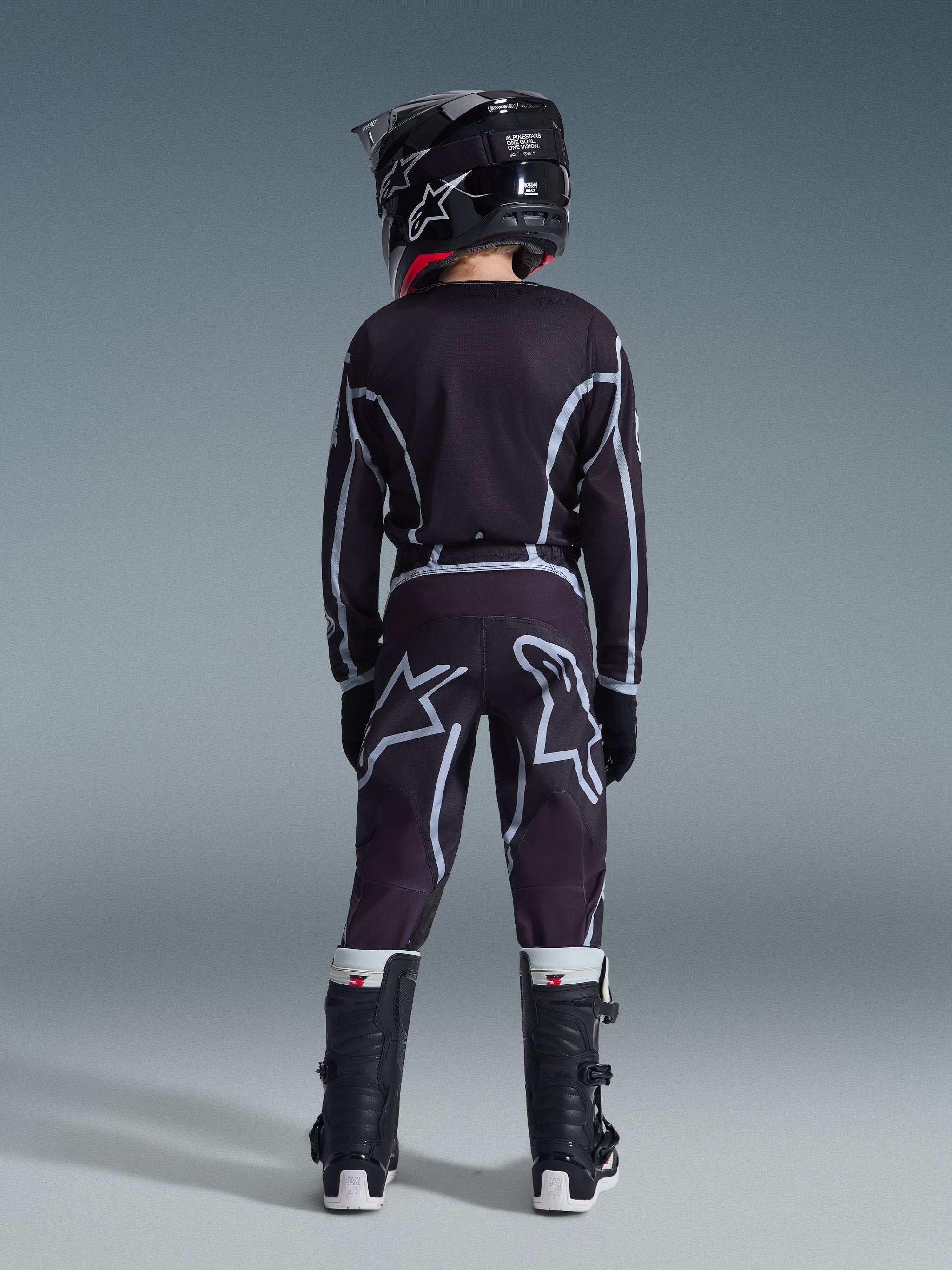 Youth motocross rider seen from behind wearing the 2026 Youth Fluid Apex Jersey in Black and Gray, matching motocross pants with large white logo graphics, black helmet, and black and white Tech Series boots, complete performance gear set for off-road riding.