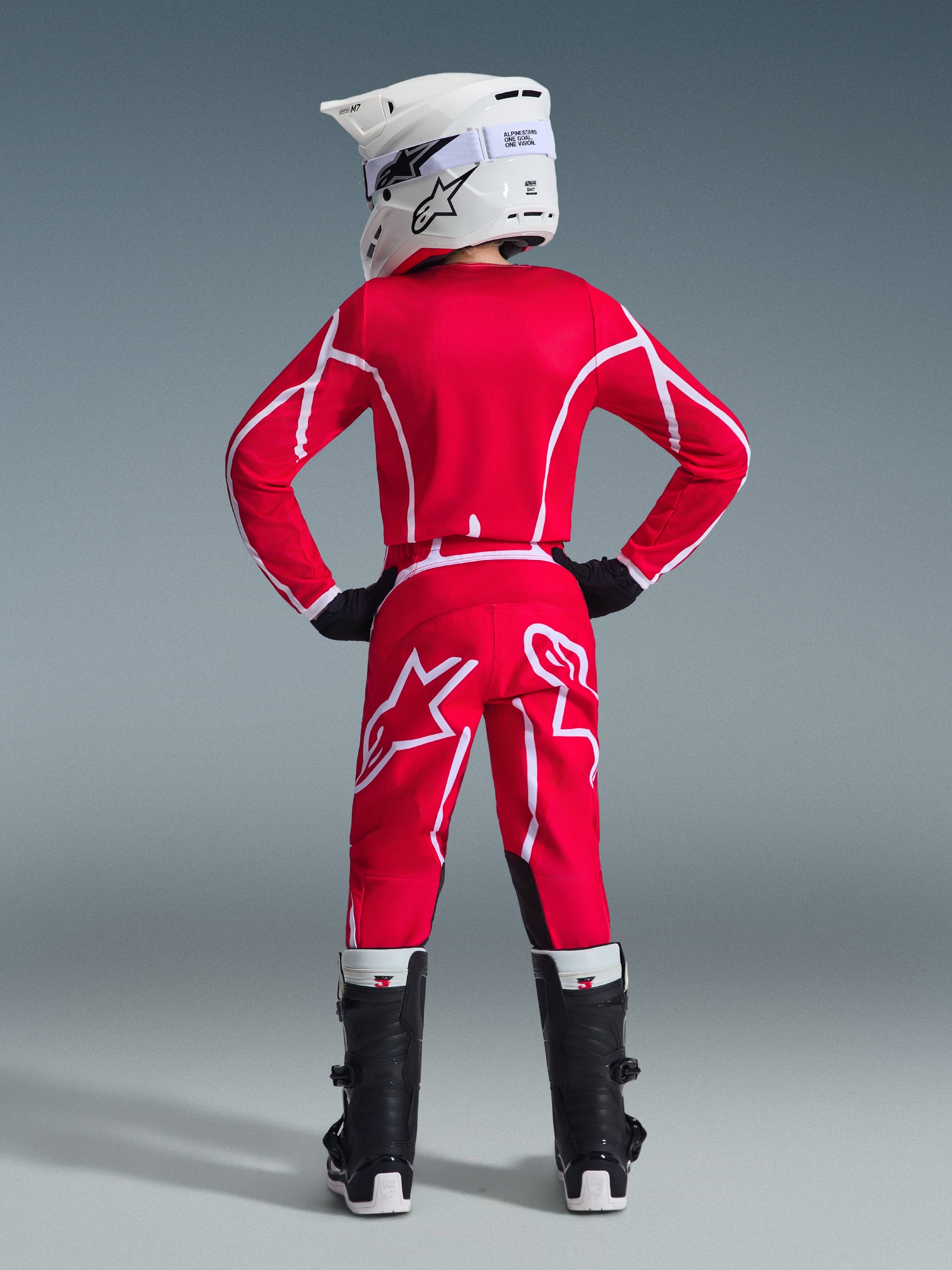 Rear view of a youth motocross rider wearing the Alpinestars 2026 Youth Fluid Apex Jersey in Red and White, matching pants, white helmet with goggles, and black boots, complete off-road racing gear set.