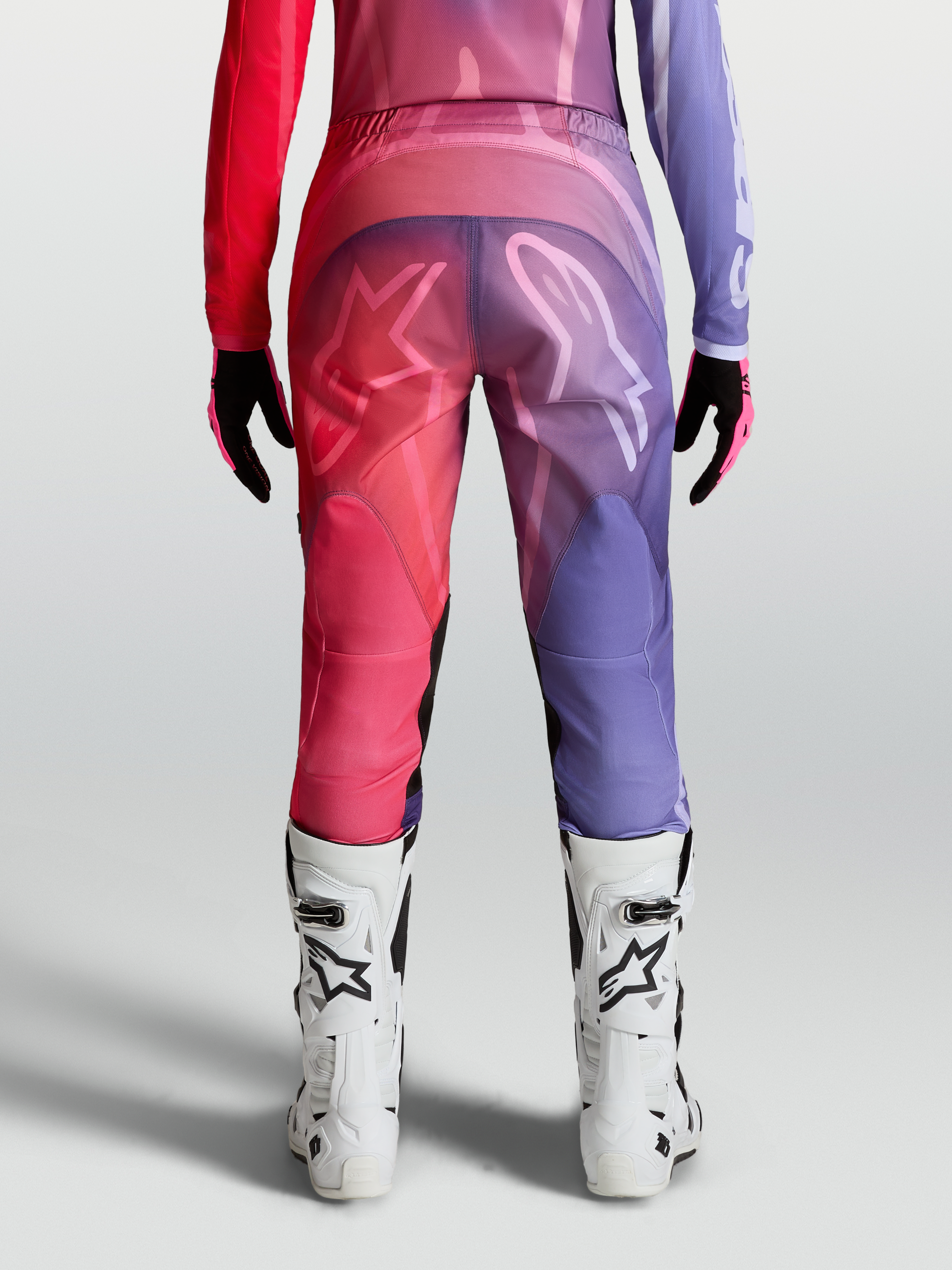 Alpinestars Stella Fluid Kepler Hose, Damen-Motocross-Hose in Lila und Rot, Rückansicht mit ergonomischem Schnitt, 600D-Polygewebe und weißen Logo-Grafiken, getragen mit weißen MX-Stiefeln.
