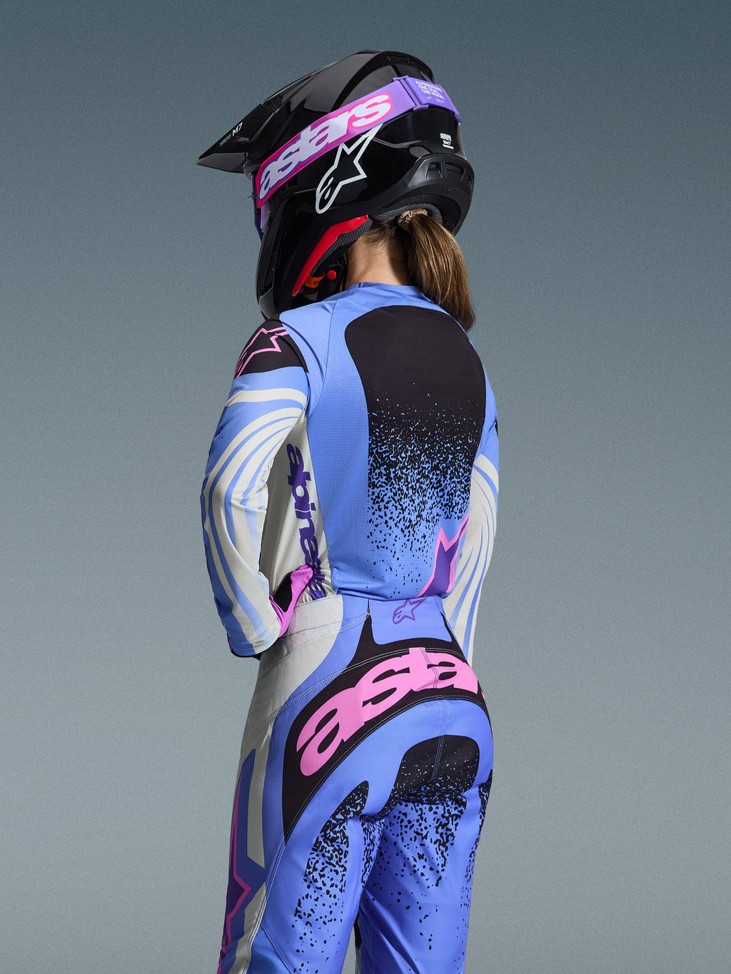 Rückansicht einer Motocross-Fahrerin im Alpinestars 2026 Stella Techstar Nomur Gear Set, Hellblau mit pinken und schwarzen Akzenten, passendes Trikot und Hose, schwarzer Helm mit pinker Brille, ergonomische Damen-Passform für Offroad-Rennsport.