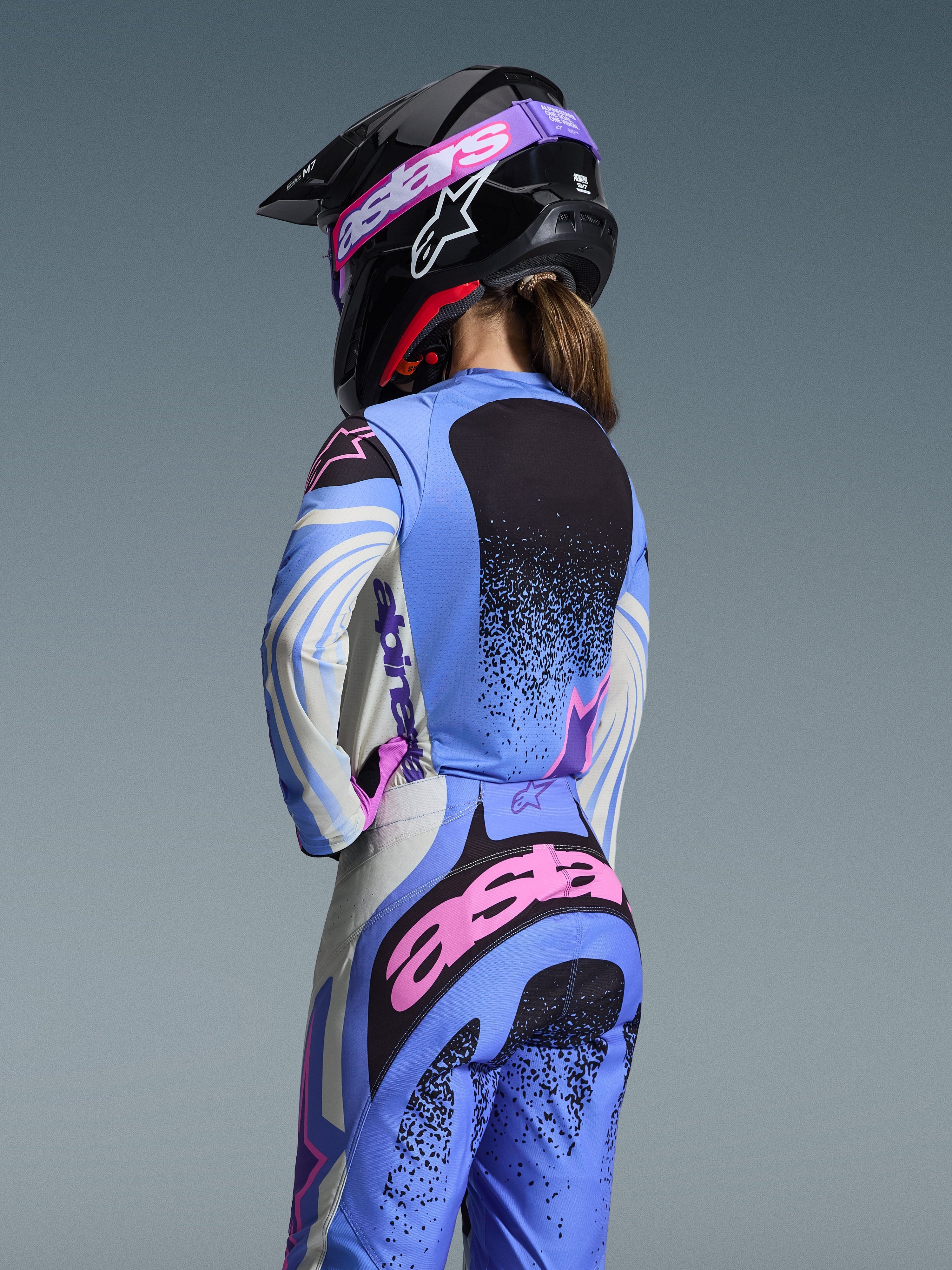 Rückansicht einer Motocross-Fahrerin im Alpinestars 2026 Stella Techstar Nomur Gear Set, Hellblau mit pinken und schwarzen Akzenten, passendes Trikot und Hose, schwarzer Helm mit pinker Brille, ergonomische Damen-Passform für Offroad-Rennsport.