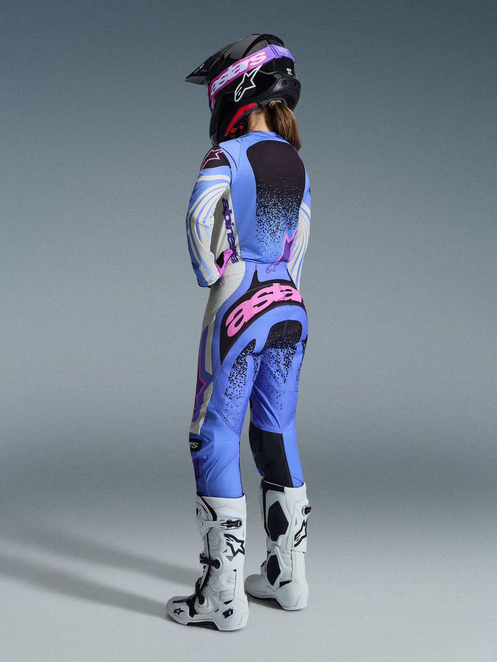 Alpinestars 2026 Stella Techstar Nomur Bekleidungsset, Rückansicht einer Motocross-Fahrerin in einem hellblauen, weißen und pinken Trikot und passender Hose mit schwarzen gesprenkelten Details, kombiniert mit einem schwarzen Helm und weißen Stiefeln, professionelle Rennbekleidung für Damen.