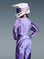 Motocross-Fahrerin im Alpinestars 2026 Stella Fluid Apex Gear Set, lila und lavendelfarbenes Trikot und Hose mit weißen Grafiken, weißer Helm mit pinkfarbener und lila Brille, spezielle Damenpassform, abgestimmte Ausrüstung für den Offroad-Rennsport