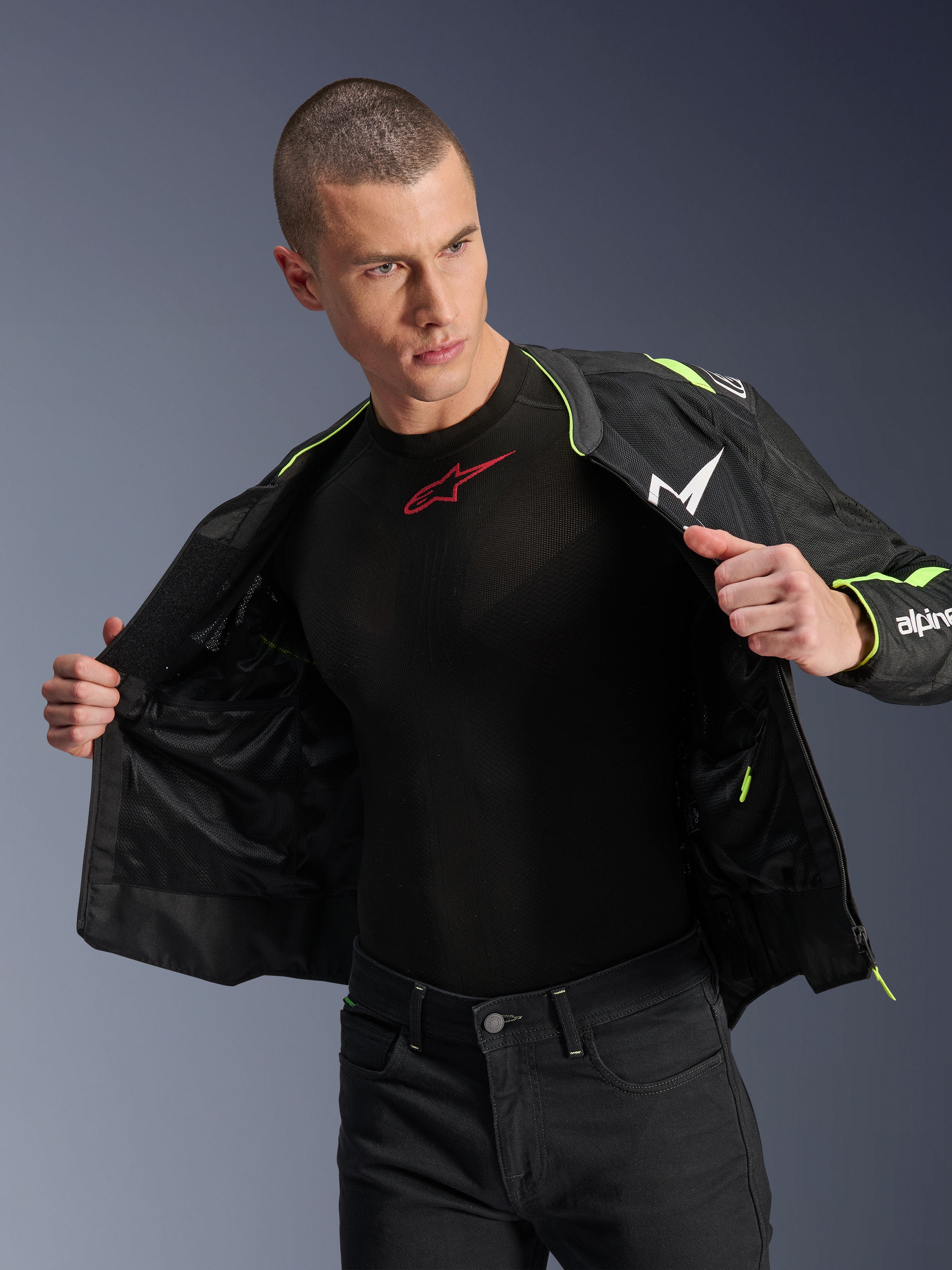 Männliches Model trägt die Alpinestars T-Sps Air V2 Jacke in Schwarz und Yellow Fluo, offen gezeigt, um die interne Mesh-Konstruktion und Taschen hervorzuheben, kombiniert mit einem schwarzen Alpinestars Base Layer und einer schwarzen Hose, urbaner Motorrad-Riding-Stil