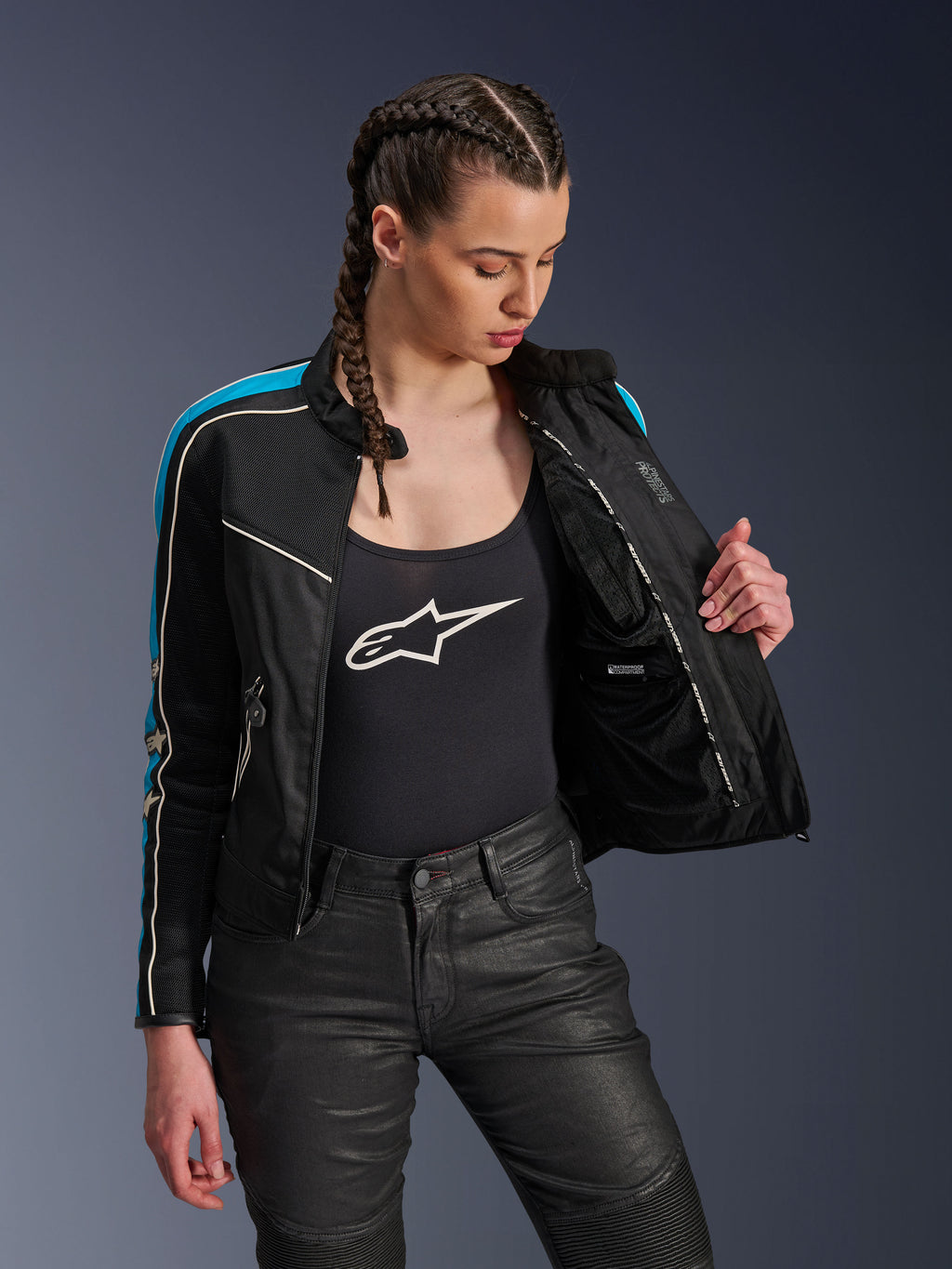 Frau trägt die Alpinestars Stella T-Dyno Air Jacke in Schwarz Hellblau Ecru, hält die Jacke offen, um das innere Mesh-Futter und die wasserdichte Tasche zu zeigen, kombiniert mit einem schwarzen Tanktop mit Logo und einer schwarzen Motorradhose, Vintage-inspirierte urbane Motorradausrüstung.