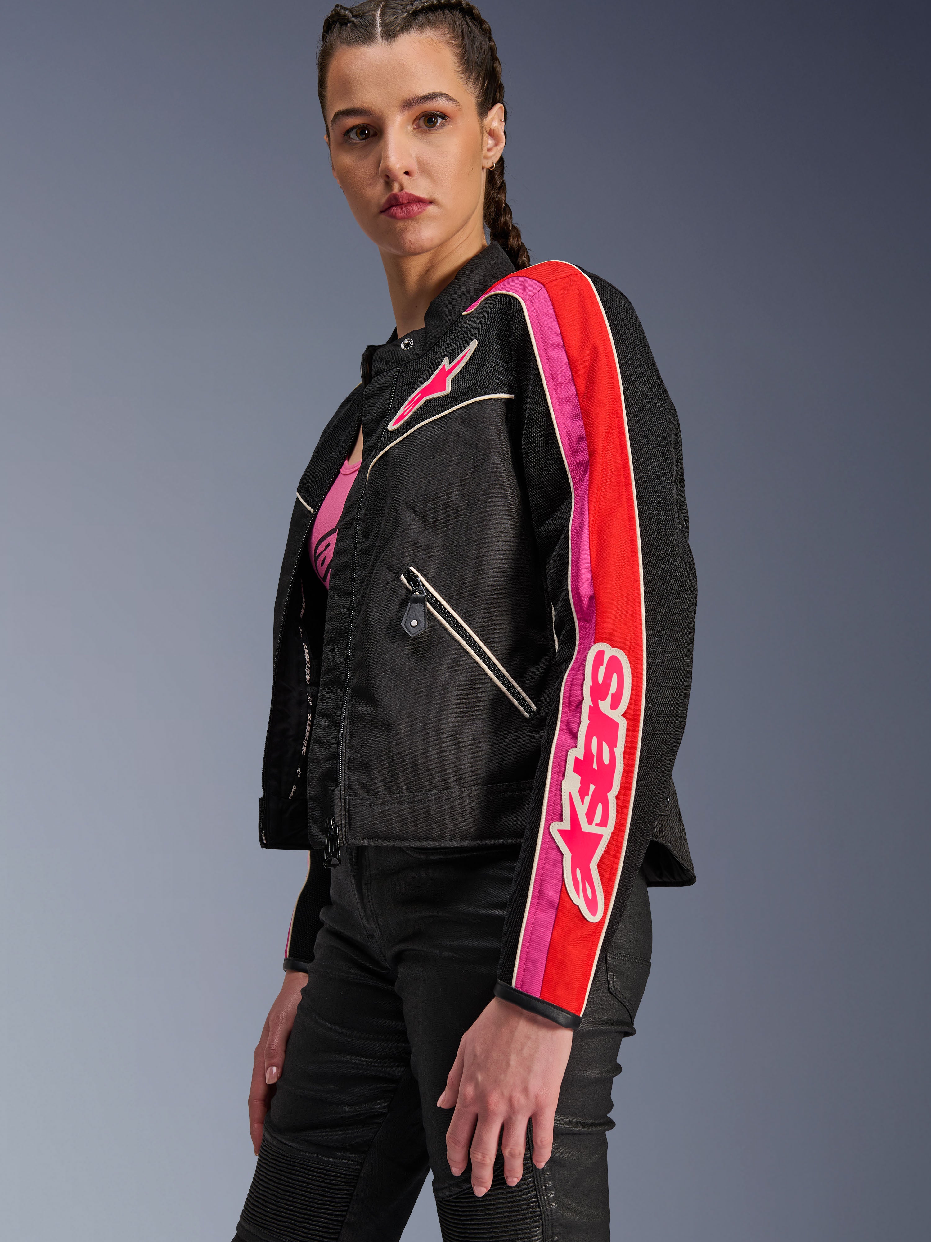 Alpinestars Stella T-Dyno Air Damenjacke, Damen-Motorradjacke, Schwarz mit leuchtend roten und fuchsiafarbenen Akzenten, getragen mit einer schwarzen Hose, mit pinkem Astars-Logo auf der Brust und am Ärmel, taillierte Biker-Passform mit Mesh-Einsätzen zur Belüftung.