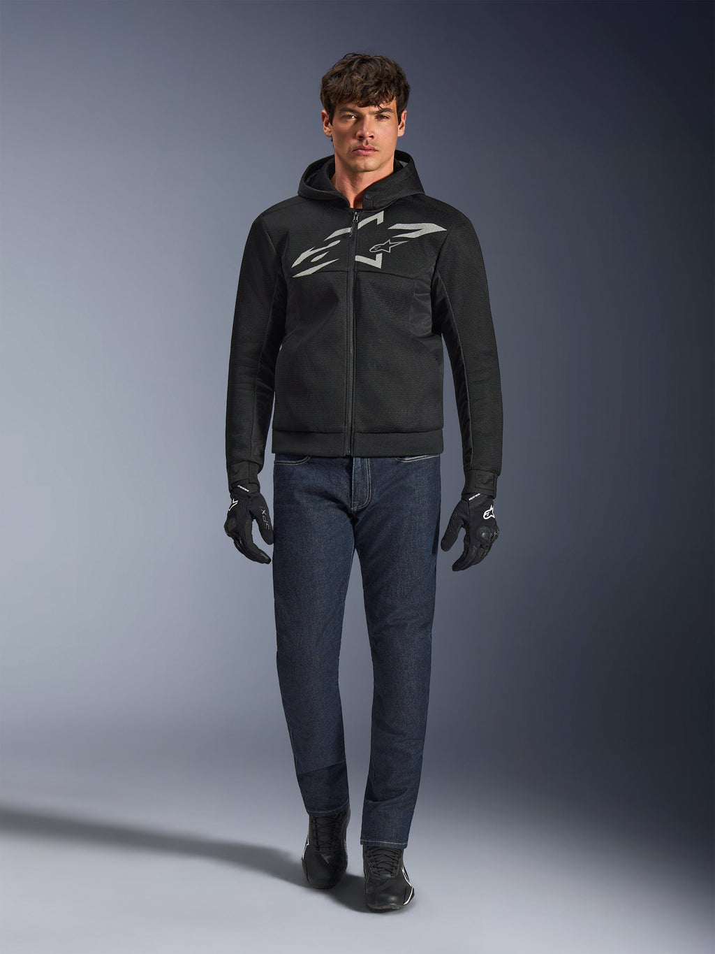 Motorcycle rider wearing the Alpinestars Chrome Superairflow Hoodie in Black and Gray, paired with dark blue jeans, black motorcycle gloves, and black shoes. The hoodie features a large stylized logo on the chest and a 3D mesh texture for high-temperature performance.