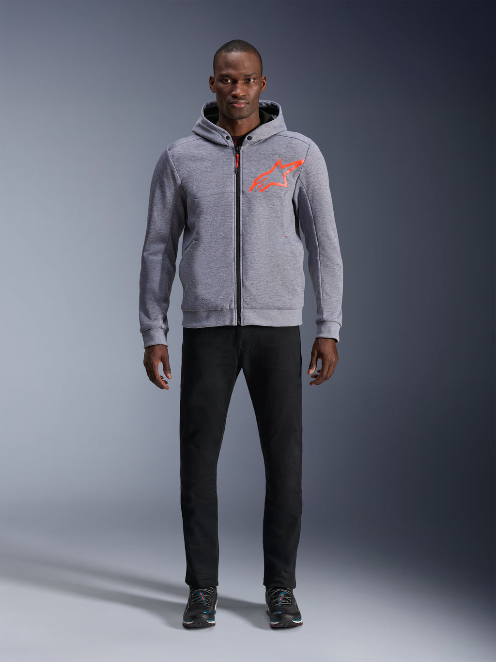 Alpinestars Chrome V2 Sport Hoodie in Gray Heather Red Fluo, hellgrauer Tech-Fleece-Hoodie mit leuchtend orangerotem Stern-Logo auf der Brust, kombiniert mit schwarzer Hose und dunklen Sneakern, lässiger urbaner Motorrad-Stil