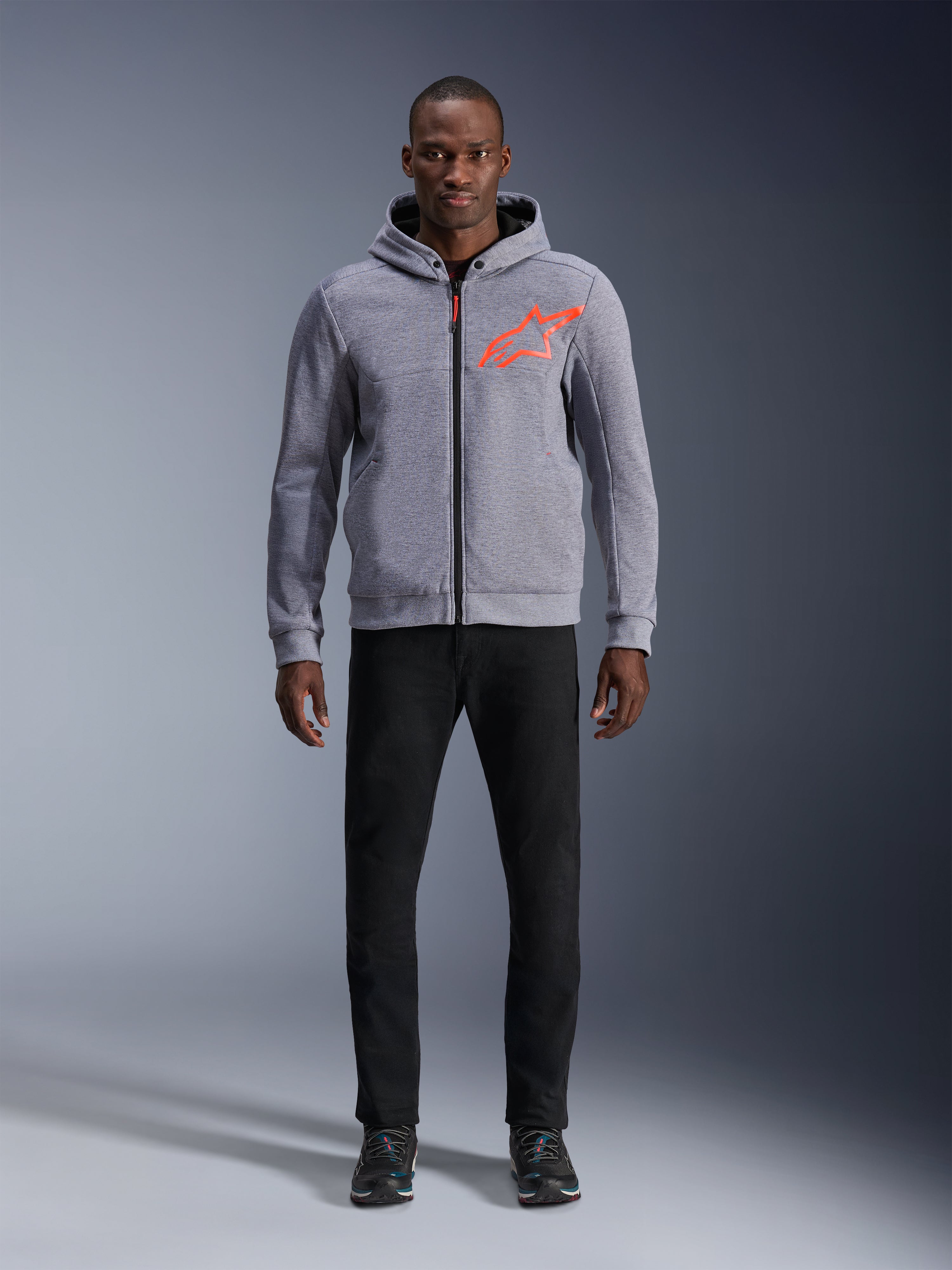 Alpinestars Chrome V2 Sport Hoodie in Gray Heather Red Fluo, hellgrauer Tech-Fleece-Hoodie mit leuchtend orangerotem Stern-Logo auf der Brust, kombiniert mit schwarzer Hose und dunklen Sneakern, lässiger urbaner Motorrad-Stil