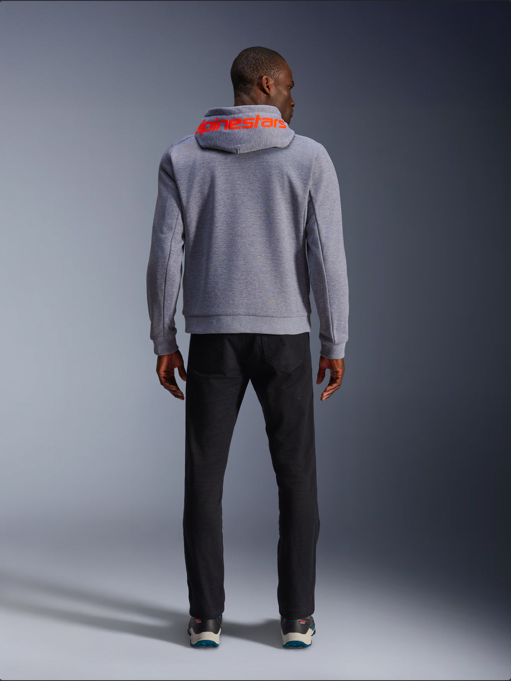 Alpinestars Chrome V2 Sport Hoodie, Grau meliert mit Red Fluo Akzenten, Rückansicht mit rotem Alpinestars-Branding auf der Kapuze, kombiniert mit schwarzer Hose, lässiger Urban-Riding-Stil mit schützender Tech-Fleece-Konstruktion