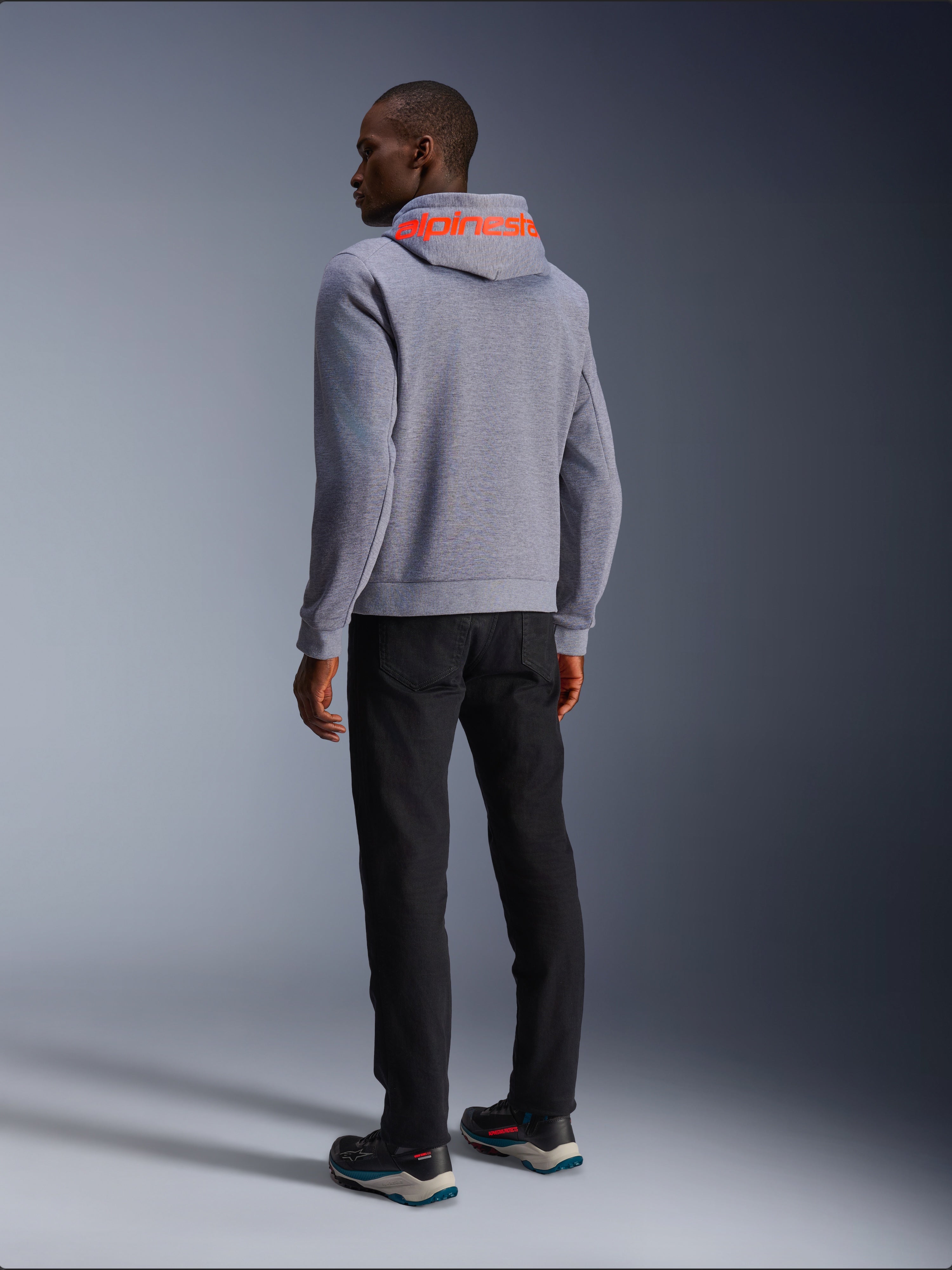 Alpinestars Chrome V2 Sport Hoodie in Gray Heather, Rückansicht mit großem Alpinestars-Schriftzug in Red Fluo auf der Kapuze, getragen von einem Model mit schwarzer Hose und dunklen Motorradschuhen, lässige urbane Motorradbekleidung