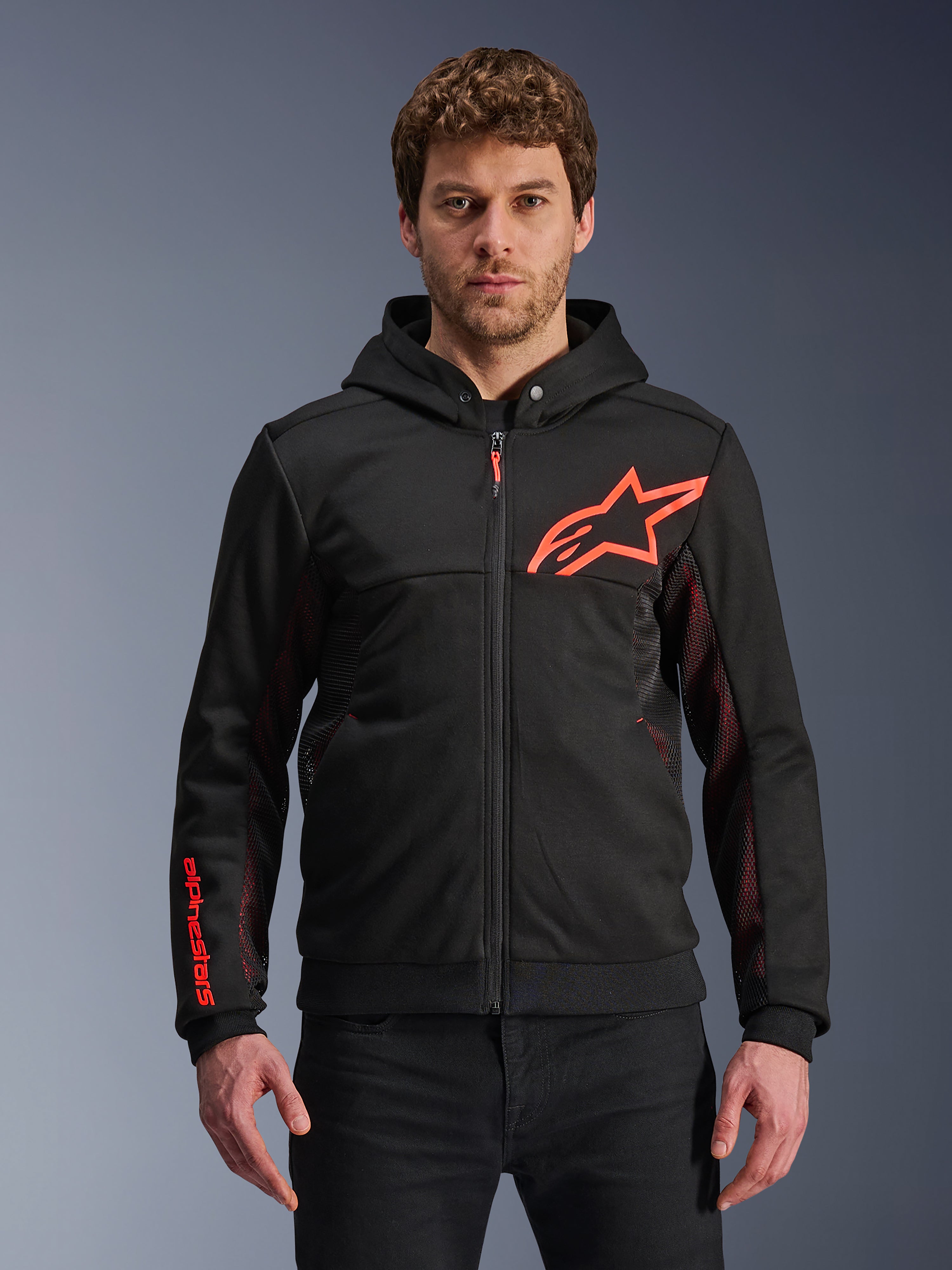 Alpinestars Chrome Air Sport Hoodie, schwarze Kapuzenjacke mit Reißverschluss, rotes Fluo-Stern-Logo und Akzente an den Ärmeln, rote Mesh-Seitenpaneele zur Belüftung, lässige urbane Motorradbekleidung, getragen von einem Model