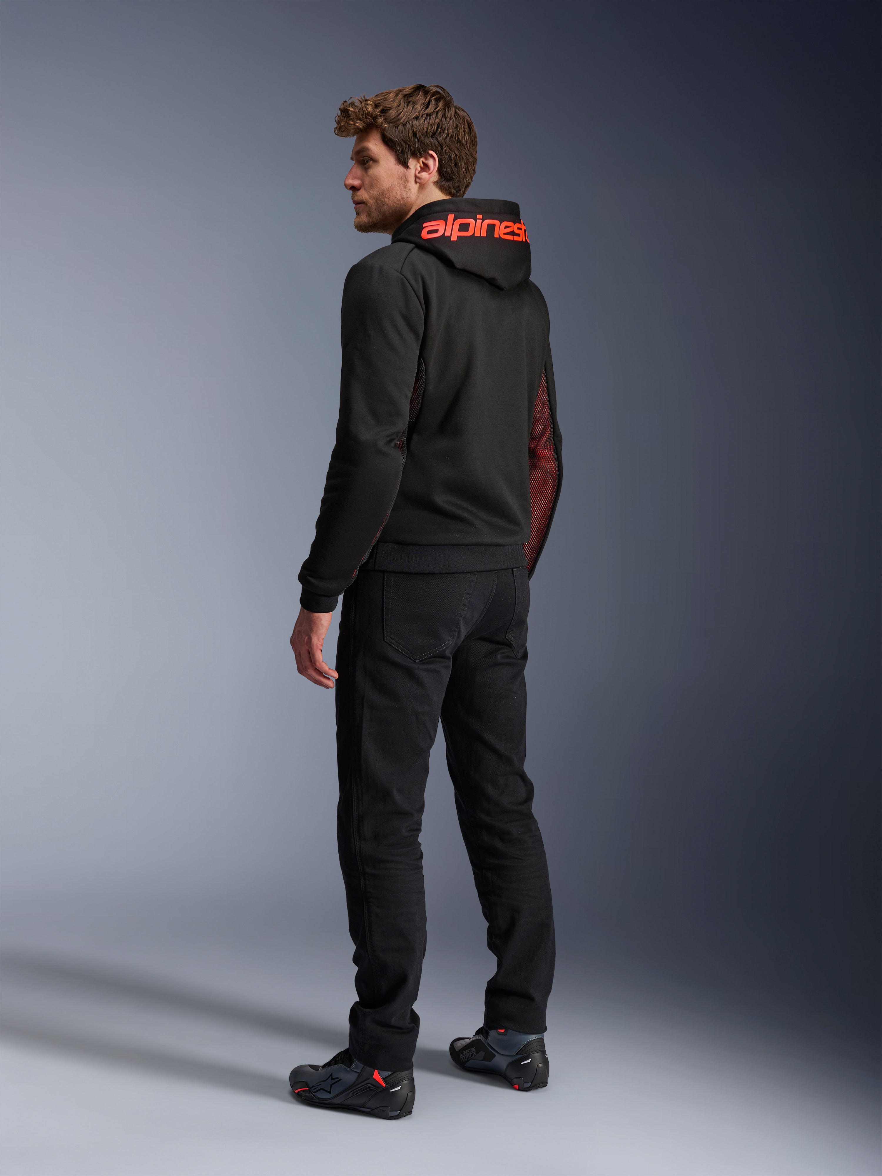 Alpinestars Chrome Air Sport Hoodie, urbaner Motorrad-Hoodie, Schwarz und Fluo-Rot, Rückansicht mit rotem Alpinestars-Branding auf der Kapuze und Mesh-Seiteneinsätzen, getragen mit schwarzer Motorradhose und technischem Schuhwerk
