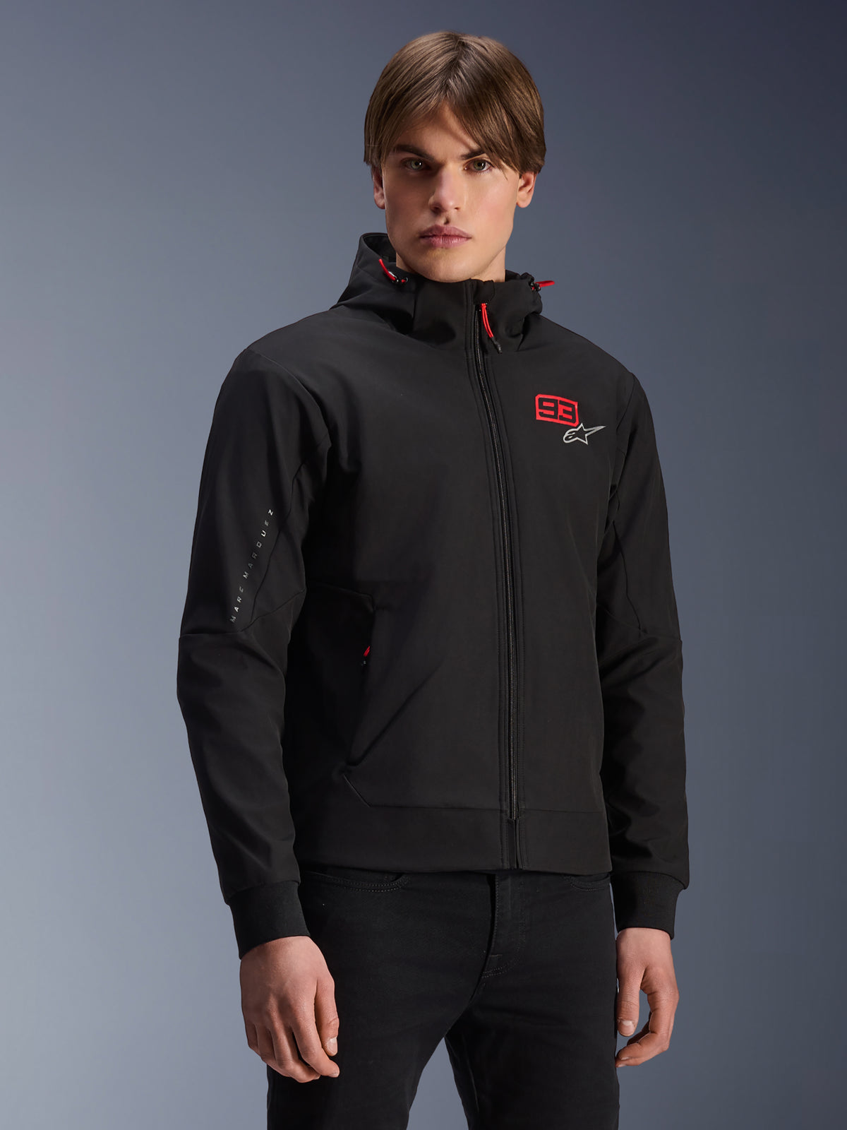 MM93 Radium Tech Hoodie, schwarze urbane Motorrad-Kapuzenjacke mit Reißverschluss, rote 93 und weißes Alpinestars-Logo auf der Brust, von einem Model mit schwarzer Hose getragen, abnehmbare Kapuze mit roten Kordelzügen und Branding am Ärmel, lässiges vom Motorsport inspiriertes Design