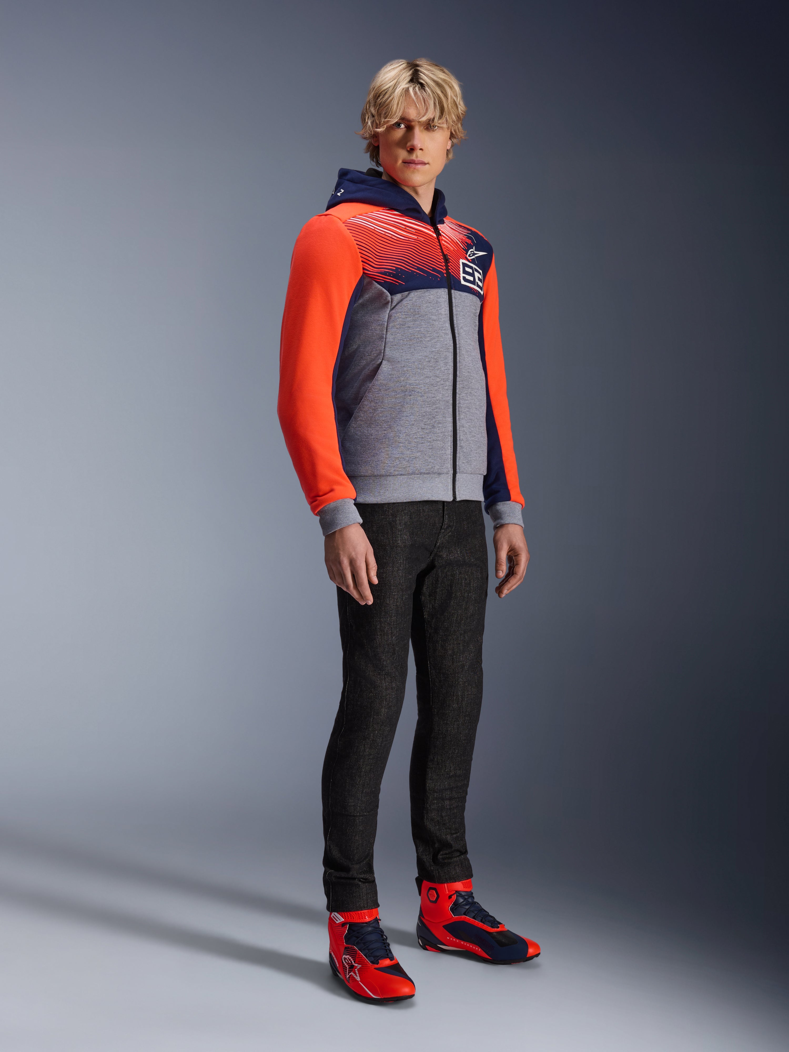 Alpinestars MM93 Chrome V2 Sport Hoodie, urban motorcycle hoodie, gray melange body with navy blue chest and bright red fluo sleeves, featuring the 93 logo on the chest, worn by a model with slim-fit black pants and coordinated red and blue high-top riding shoes, modern athletic fit for street riding