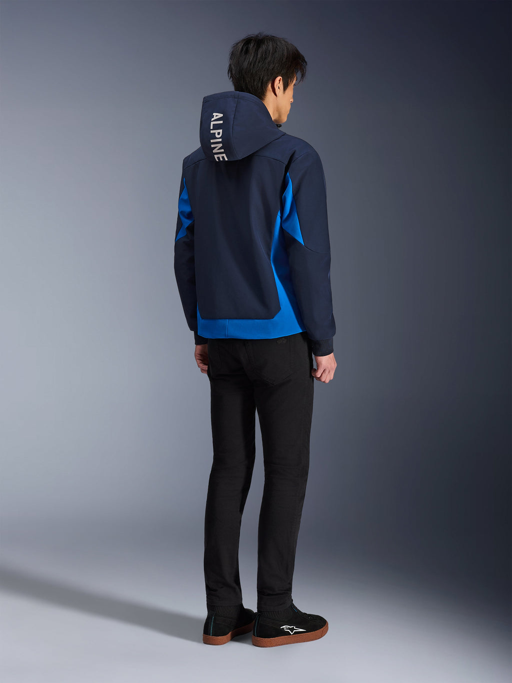 Alpinestars Radium Tech Hoodie, Night Navy und Royal Blue, Rückansicht eines Models, das den technischen Shell-Hoodie mit weißem „ALPINE“-Schriftzug auf der Kapuze trägt, kombiniert mit schwarzer Hose und Schuhen, urbane Motorradbekleidung