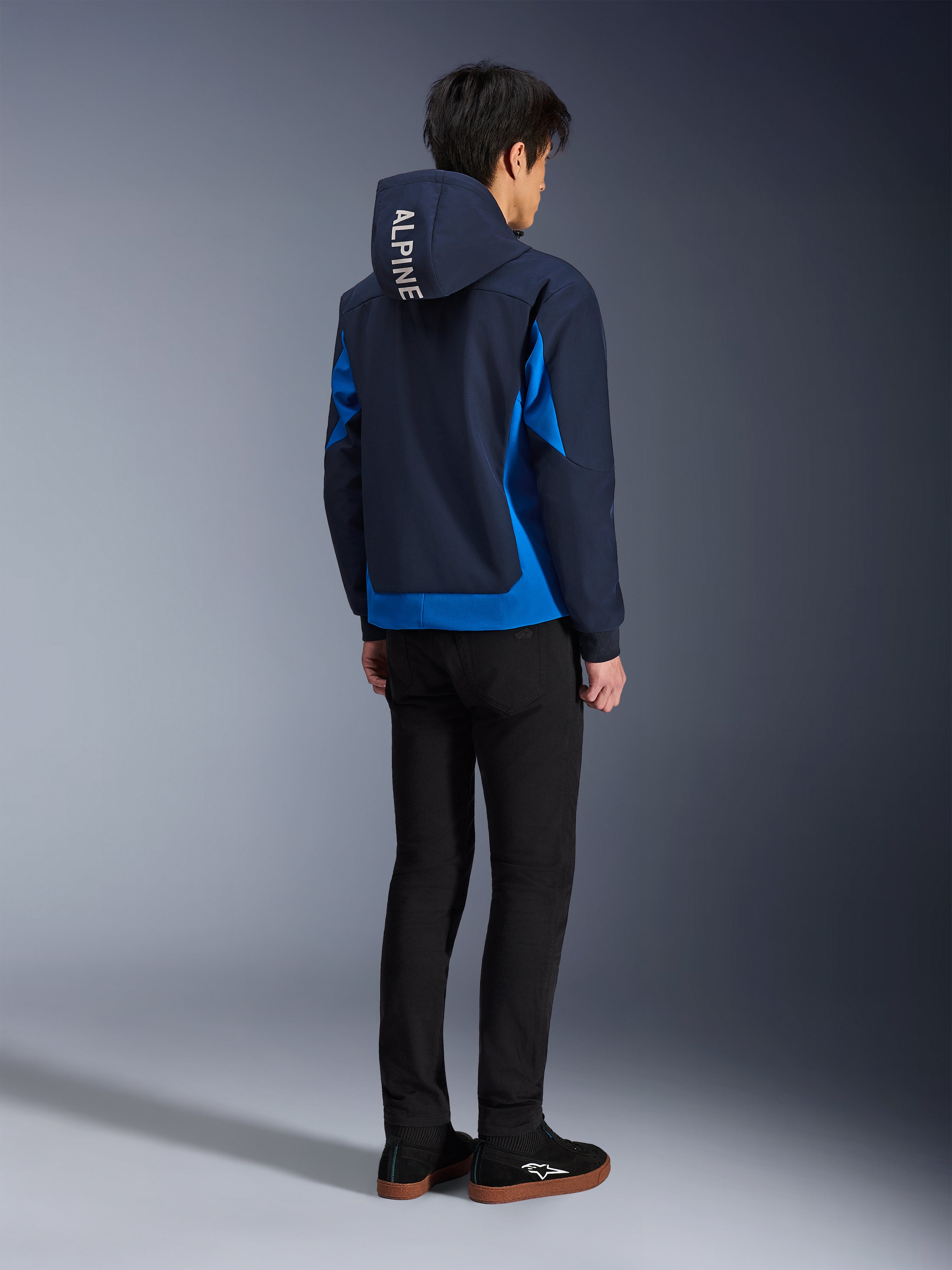 Alpinestars Radium Tech Hoodie, Night Navy und Royal Blue, Rückansicht eines Models, das den technischen Shell-Hoodie mit weißem „ALPINE“-Schriftzug auf der Kapuze trägt, kombiniert mit schwarzer Hose und Schuhen, urbane Motorradbekleidung