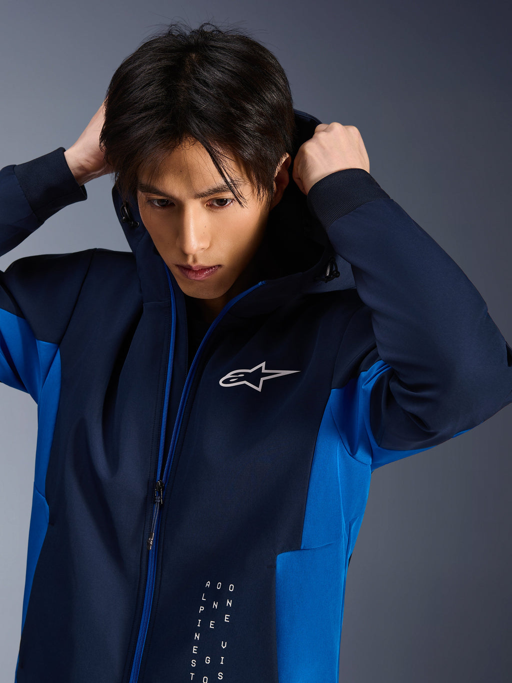 Alpinestars Radium Tech Hoodie, urbane Motorradjacke, Night Navy und Royal Blue, Modell beim Anpassen der Kapuze, mit weißem Astars-Logo auf der Brust und technischem 3L-Shell-Design mit athletischer Passform.