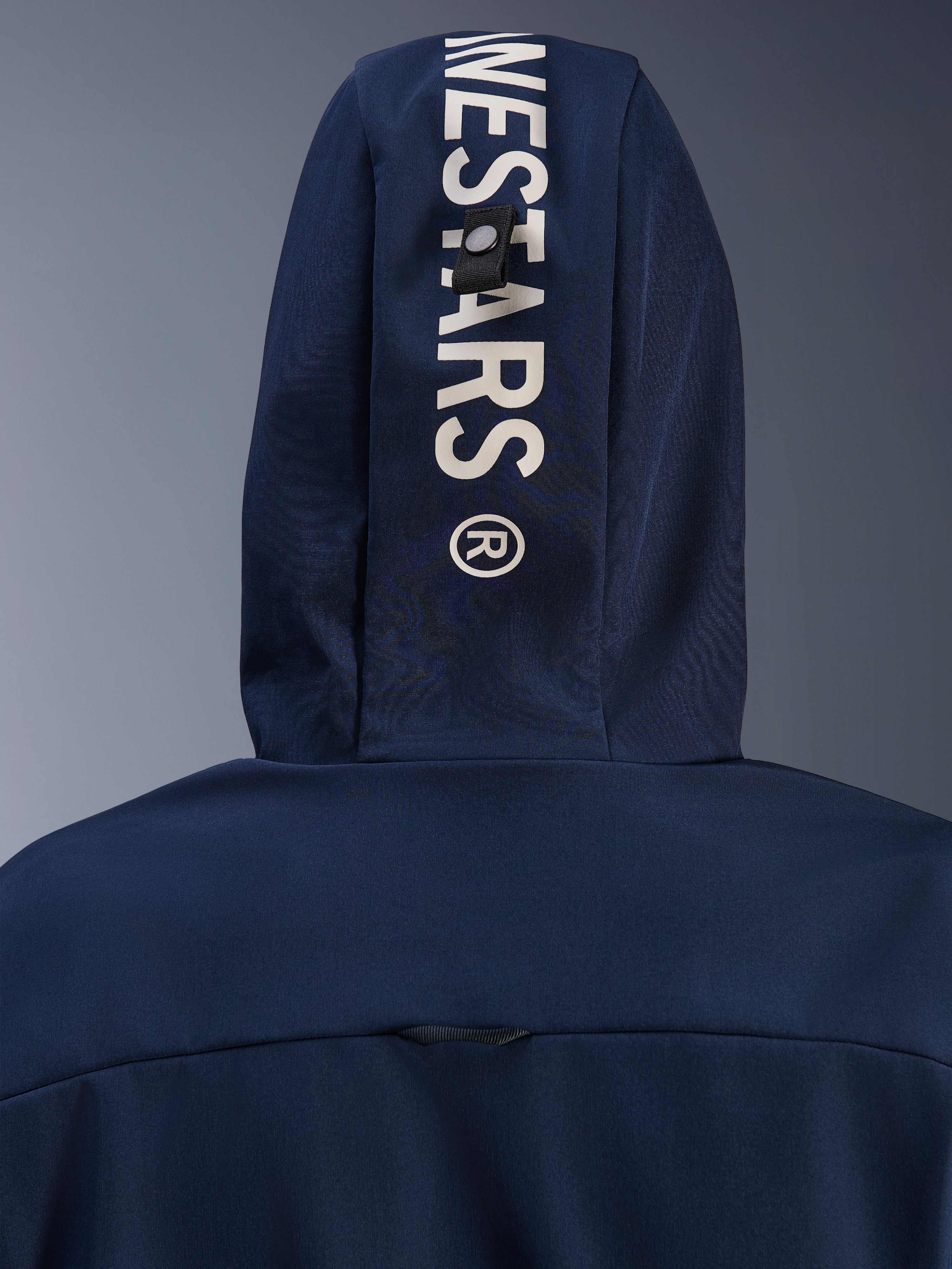 Alpinestars Radium Tech Hoodie in Night Navy Royal Blue, Nahaufnahme der Kapuzenrückseite mit vertikalem weißem Alpinestars-Branding und einem sicheren Befestigungsschlaufen-System, urbane technische Motorradbekleidung.