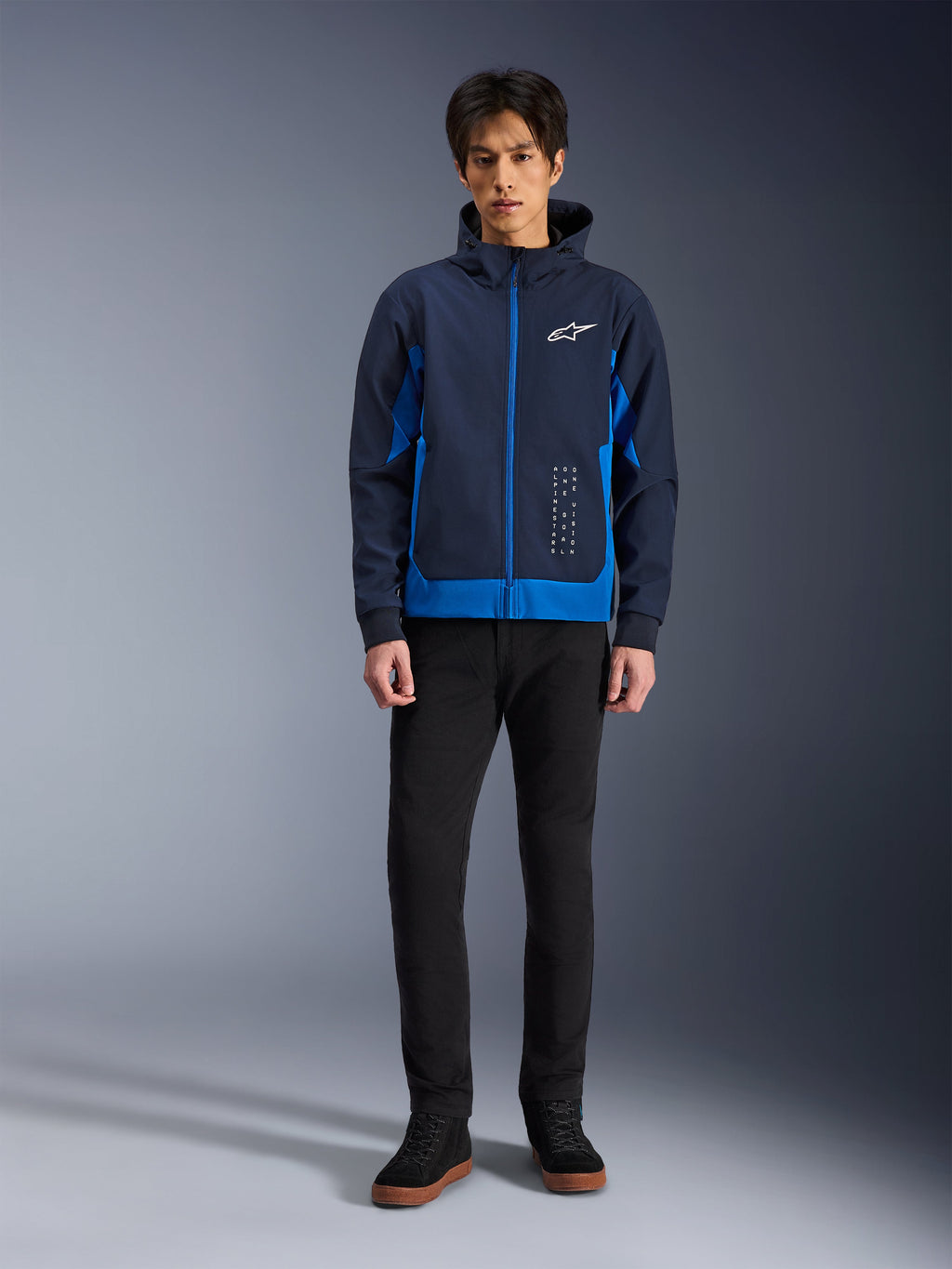 Alpinestars Radium Tech Hoodie, urbaner Motorrad-Hoodie in Night Navy und Royal Blue, winddichtes Technical-Shell, lässiges Streetwear-Design kombiniert mit schwarzer Hose und High-Top-Sneakern, mit weißem Logo-Branding auf der Brust.
