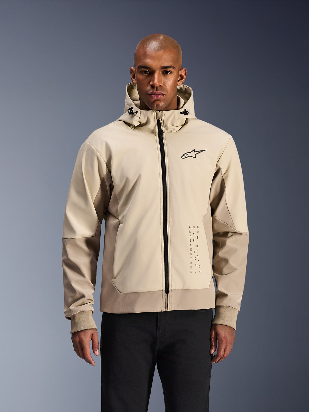 Alpinestars Radium Tech Hoodie in Mastic Aluminum, hellbeige und hellbraune urbane Motorradjacke, schwarzer Frontreißverschluss und kleines Brustlogo, präsentiert von einem Mann in schwarzer Hose, technisches winddichtes Außenmaterial mit modernem Streetwear-Styling