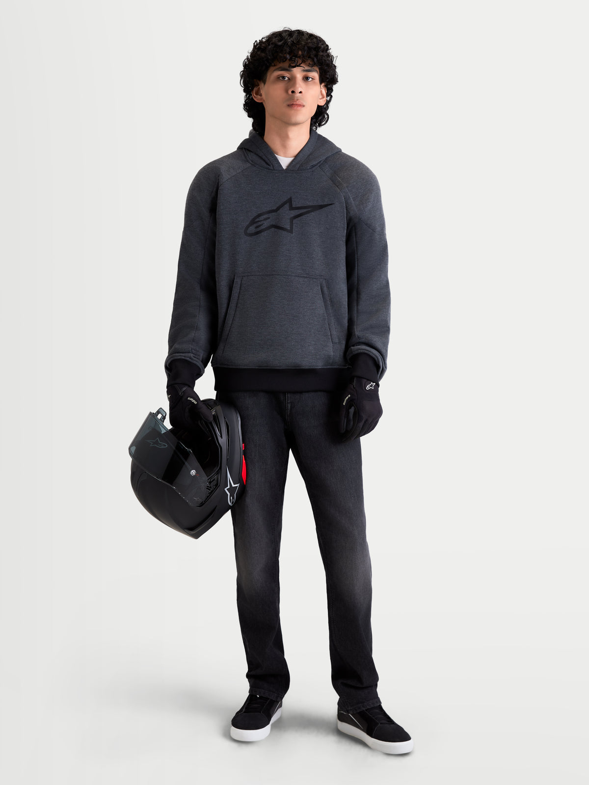 Alpinestars Ageless Kapuzenpullover, urbaner Motorrad-Kapuzenpullover für Herren in Asphalt-Melange-Grau, Frontansicht an einem Model mit schwarzen Motorradhandschuhen, das einen Helm hält, mit integriertem Schulter- und Ellbogenschutz