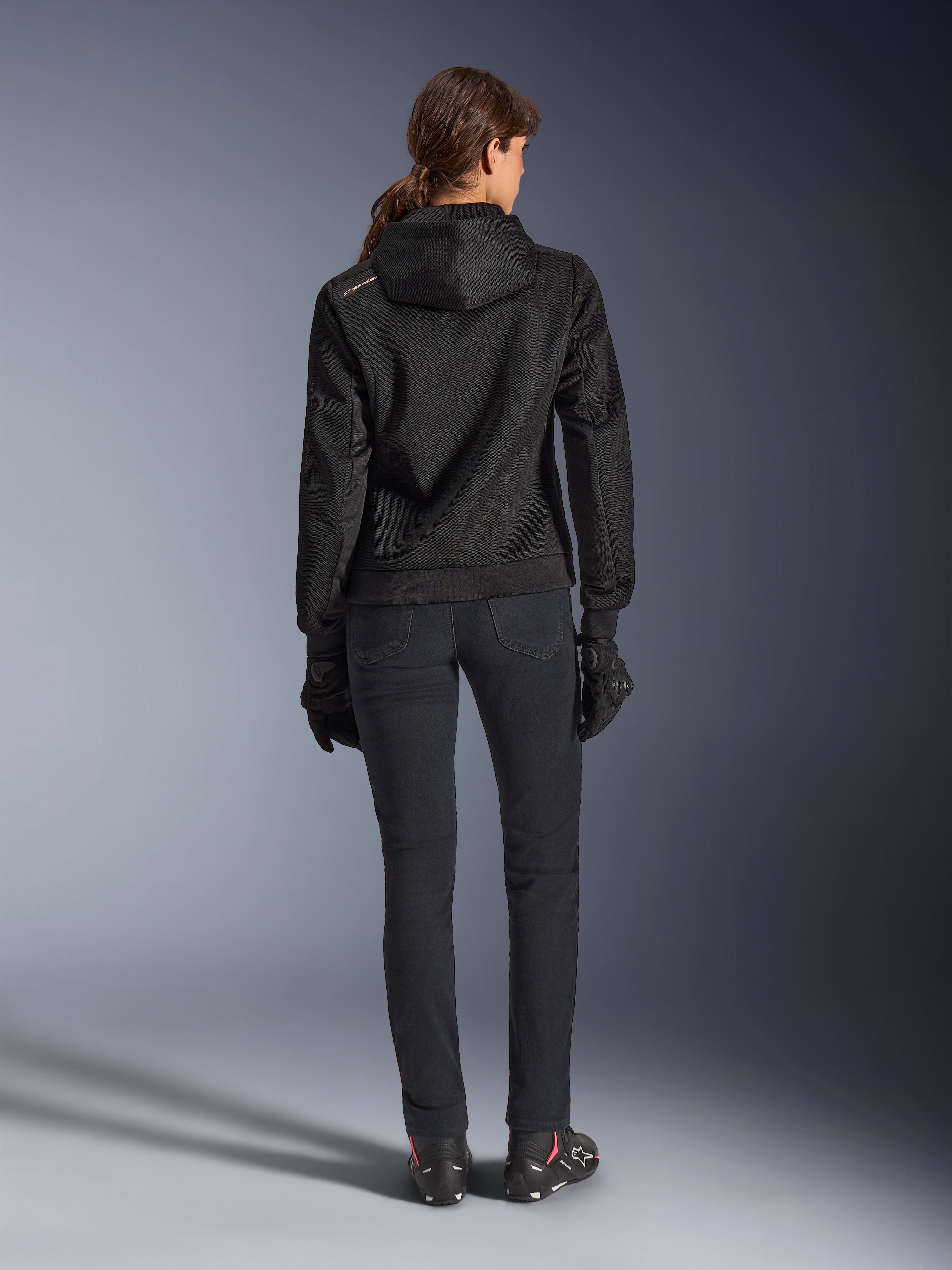 Alpinestars Stella Chrome Superairflow Hoodie, women's technical motorcycle hoodie, black 3D mesh with small orange logo on the shoulder, rear view, worn with black riding pants, black gloves and boots, designed for urban sport riding