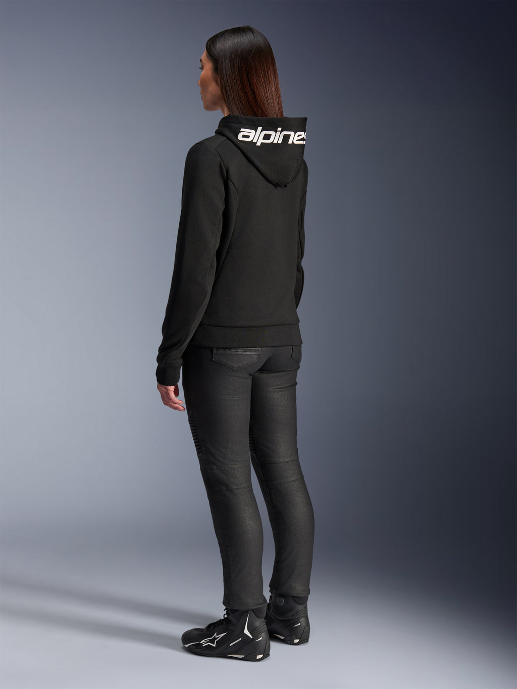 Frau trägt Alpinestars Stella Chrome V2 Sport Hoodie, Rückansicht, schwarzer Motorrad-Hoodie mit weißem Alpinestars-Logo auf der Kapuze, kombiniert mit schwarzer technischer Fahrhose und schwarzen Motorradschuhen, lässige urbane Pendlerausrüstung