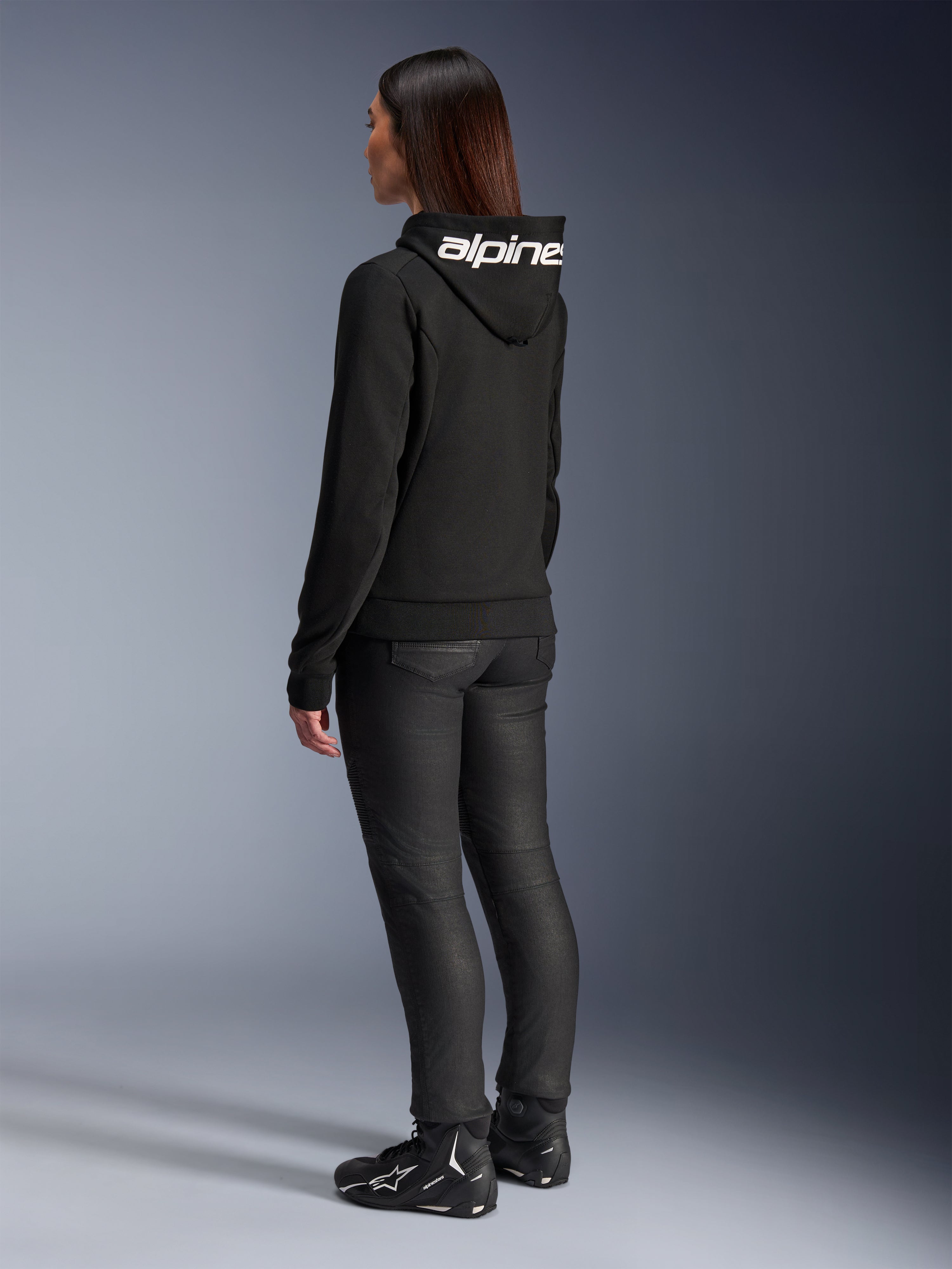 Frau trägt Alpinestars Stella Chrome V2 Sport Hoodie, Rückansicht, schwarzer Motorrad-Hoodie mit weißem Alpinestars-Logo auf der Kapuze, kombiniert mit schwarzer technischer Fahrhose und schwarzen Motorradschuhen, lässige urbane Pendlerausrüstung