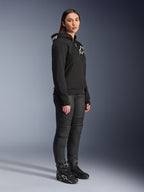 Frau im Alpinestars Stella Chrome V2 Sport Hoodie in Schwarz mit weißem Alpinestars-Branding, kombiniert mit schwarzer Motorradhose und Stiefeln, urbane Motorradausrüstung für Damen mit lässiger, sportlicher Passform