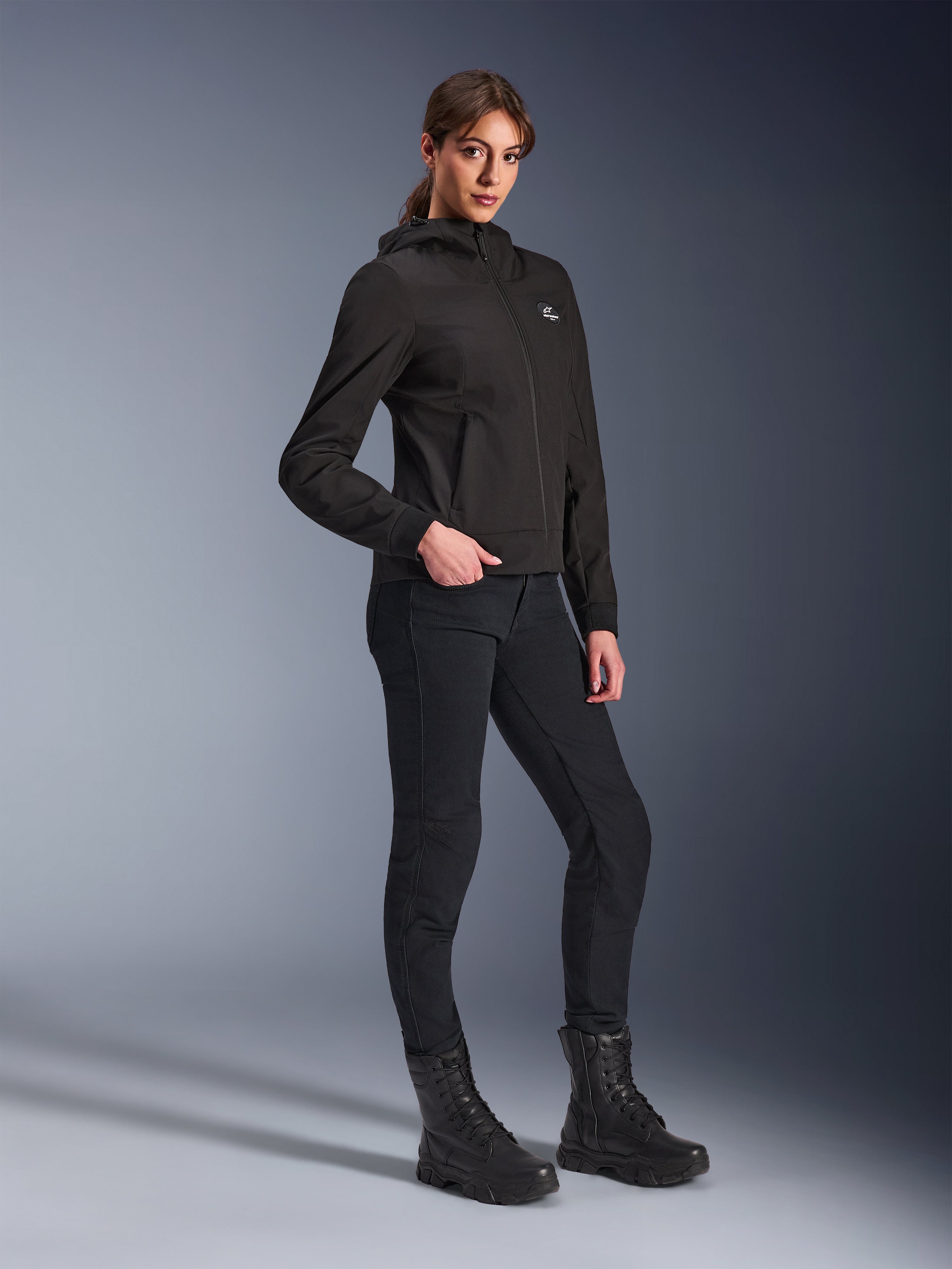 Alpinestars Stella Radium Tech Hoodie, Black urban motorcycle hoodie for women, worn with black riding pants and tactical-style boots, slim athletic fit, windproof 3-layer tech shell, minimalist design with small chest logo, full-length studio shot