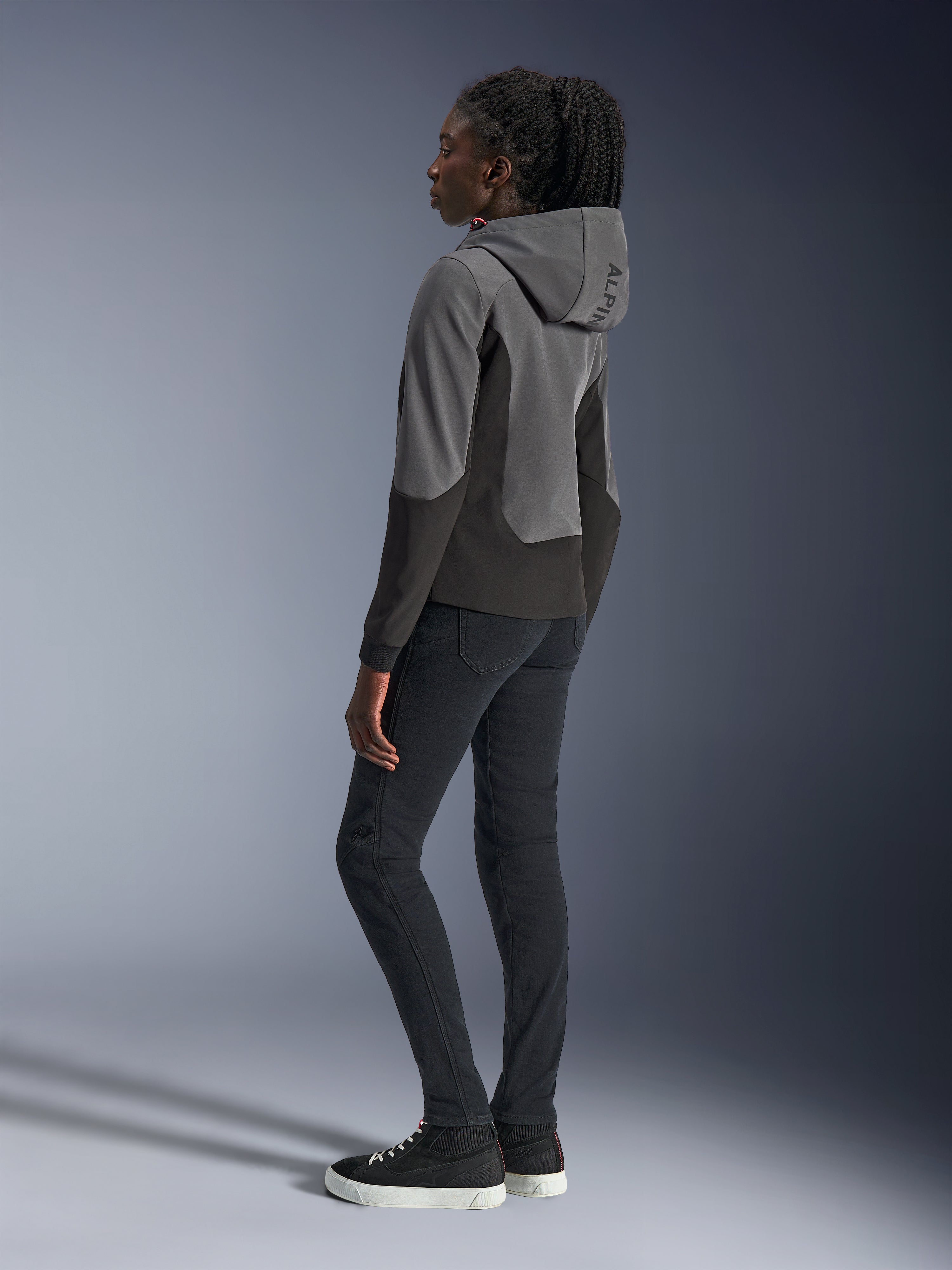 Woman wearing Alpinestars Stella Radium Tech Hoodie in Deep Gray and Black, urban motorcycle apparel, back view showing hood with Alpinestars branding, styled with black slim-fit pants and high-top sneakers