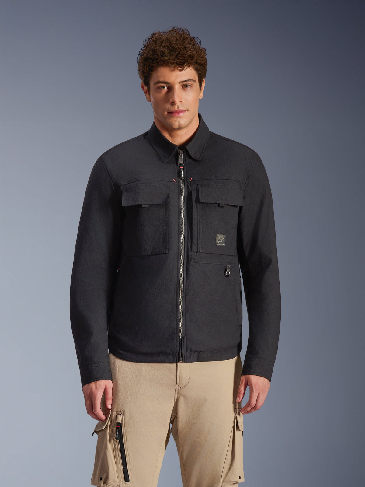 Alpinestars Bryon Water Resistant Overshirt, Schwarz, urbane Motorradjacke mit Workwear-inspiriertem Styling, zwei große Brust-Cargo-Taschen, durchgehender Reißverschluss, aus gewachstem Canvas, getragen von einem Modell mit beigefarbener Cargohose für einen modernen Utility-Look.