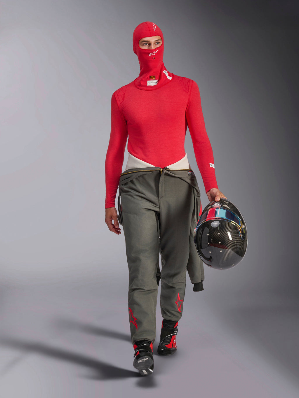 Alpinestars ZX Evo V3 Sturmhaube in Rot, getragen von einem Rennfahrer mit passendem roten langärmeligen Base-Layer und grauer Rennhose, hält einen Carbonfaser-Helm, professionelle flammhemmende Autorenn-Ausrüstung