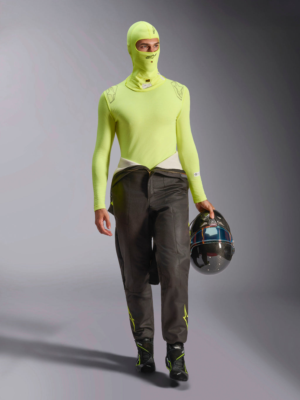 Männlicher Rennfahrer mit Alpinestars ZX Evo V3 Sturmhaube in Yellow Fluo, passendes langärmliges Base-Layer-Oberteil, dunkle Rennhose und schwarze Rennstiefel, hält einen Carbonfaser-Helm, professionelle Automobil-Rennausrüstung