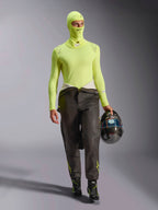 Männlicher Rennfahrer mit Alpinestars ZX Evo V3 Sturmhaube in Yellow Fluo, passendes langärmliges Base-Layer-Oberteil, dunkle Rennhose und schwarze Rennstiefel, hält einen Carbonfaser-Helm, professionelle Automobil-Rennausrüstung