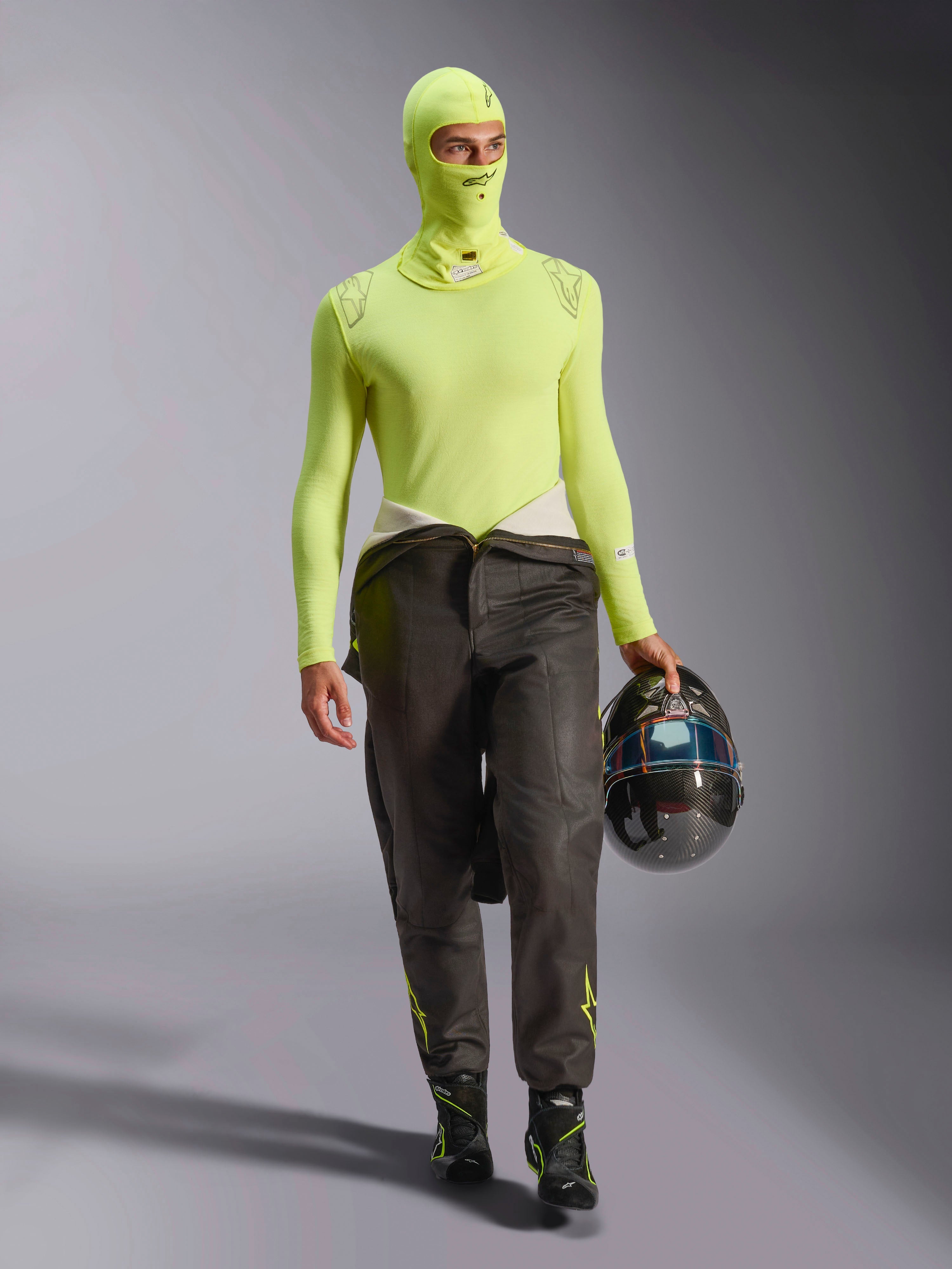 Männlicher Rennfahrer mit Alpinestars ZX Evo V3 Sturmhaube in Yellow Fluo, passendes langärmliges Base-Layer-Oberteil, dunkle Rennhose und schwarze Rennstiefel, hält einen Carbonfaser-Helm, professionelle Automobil-Rennausrüstung
