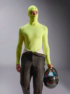 Alpinestars ZX Evo V3 Sturmhaube, Motorsport-Unterwäsche, Yellow Fluo, getragen mit passendem Langarm-Oberteil und Rennanzughose, Modell hält einen Carbon-Helm mit irisierendem Visier, professionelle flammhemmende Motorsport-Ausrüstung