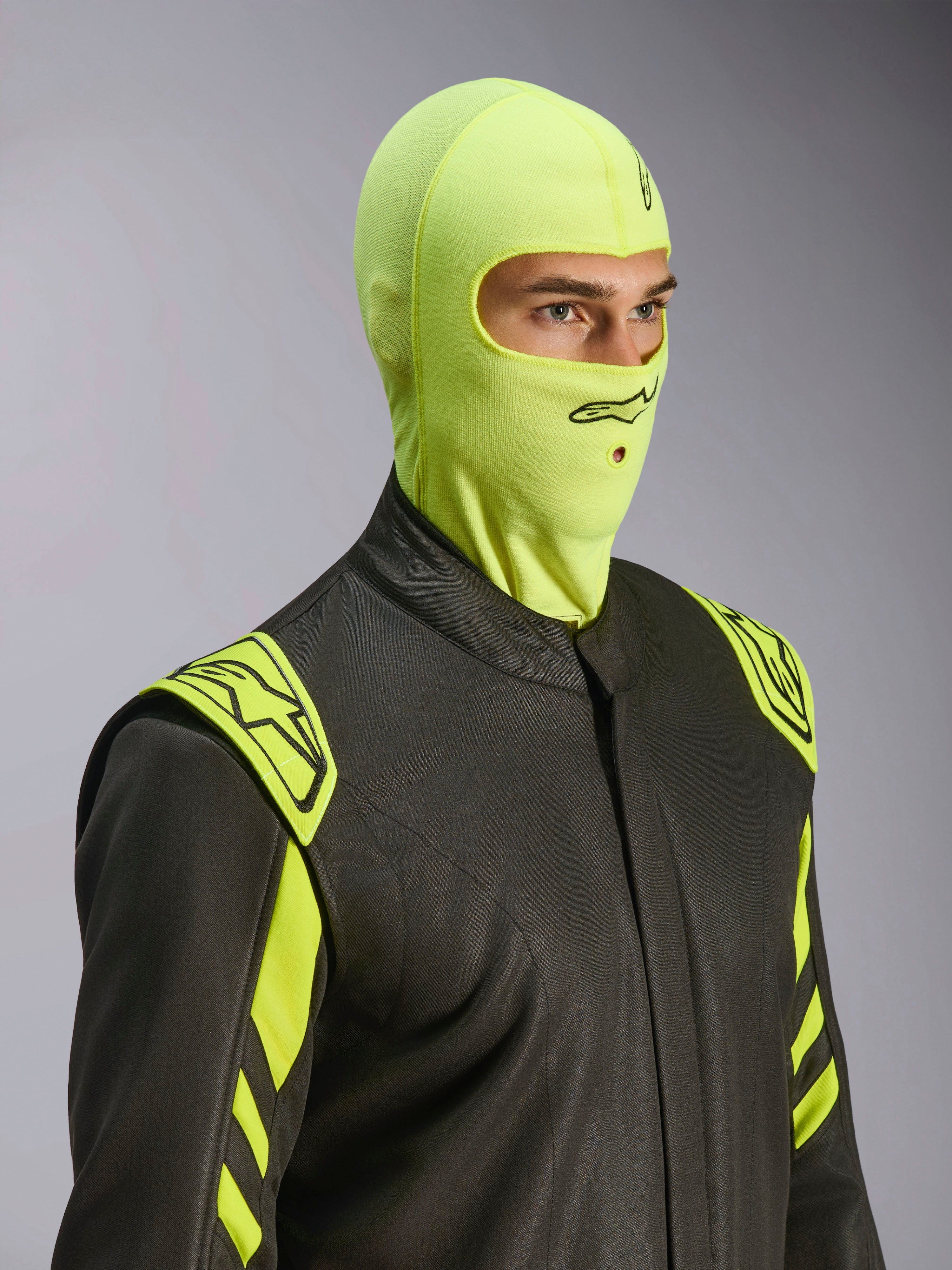 Männliches Model in der Alpinestars ZX Evo V3 Sturmhaube in Yellow Fluo, flammenhemmende Rennsport-Kopfhaube, kombiniert mit einem dunkelgrauen Autorennanzug mit neongelben Akzenten, mit schwarzem Alpinestars-Logo und Mundöffnung für Trinkschlauch, professionelle Motorsport-Schutzausrüstung.