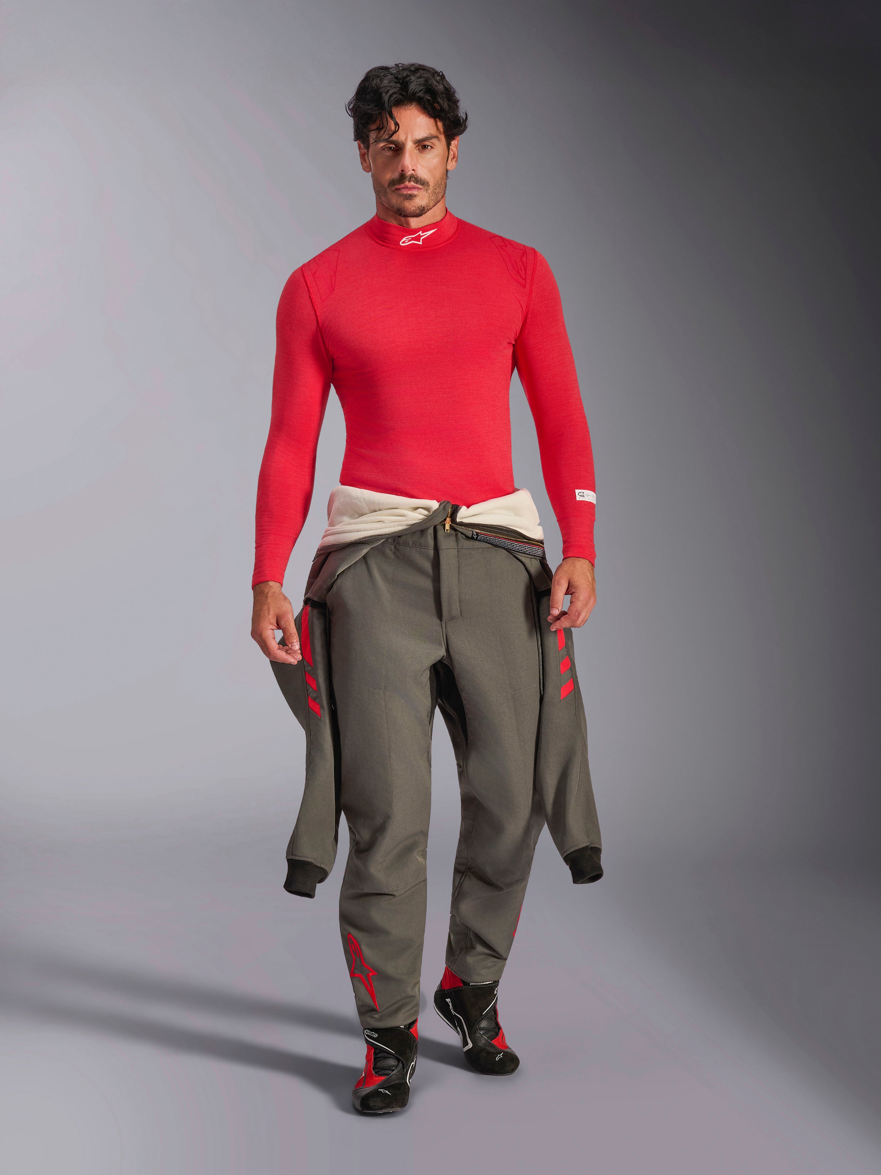 Alpinestars ZX Evo V3 Langarm-Top, rote Auto-Funktionswäsche, getragen von einem Model mit einem grauen Rennanzug, der um die Taille gebunden ist, und Rennschuhen, Stehkragen mit weißem Alpinestars-Logo, leichte und flammhemmende professionelle Motorsport-Ausrüstung.
