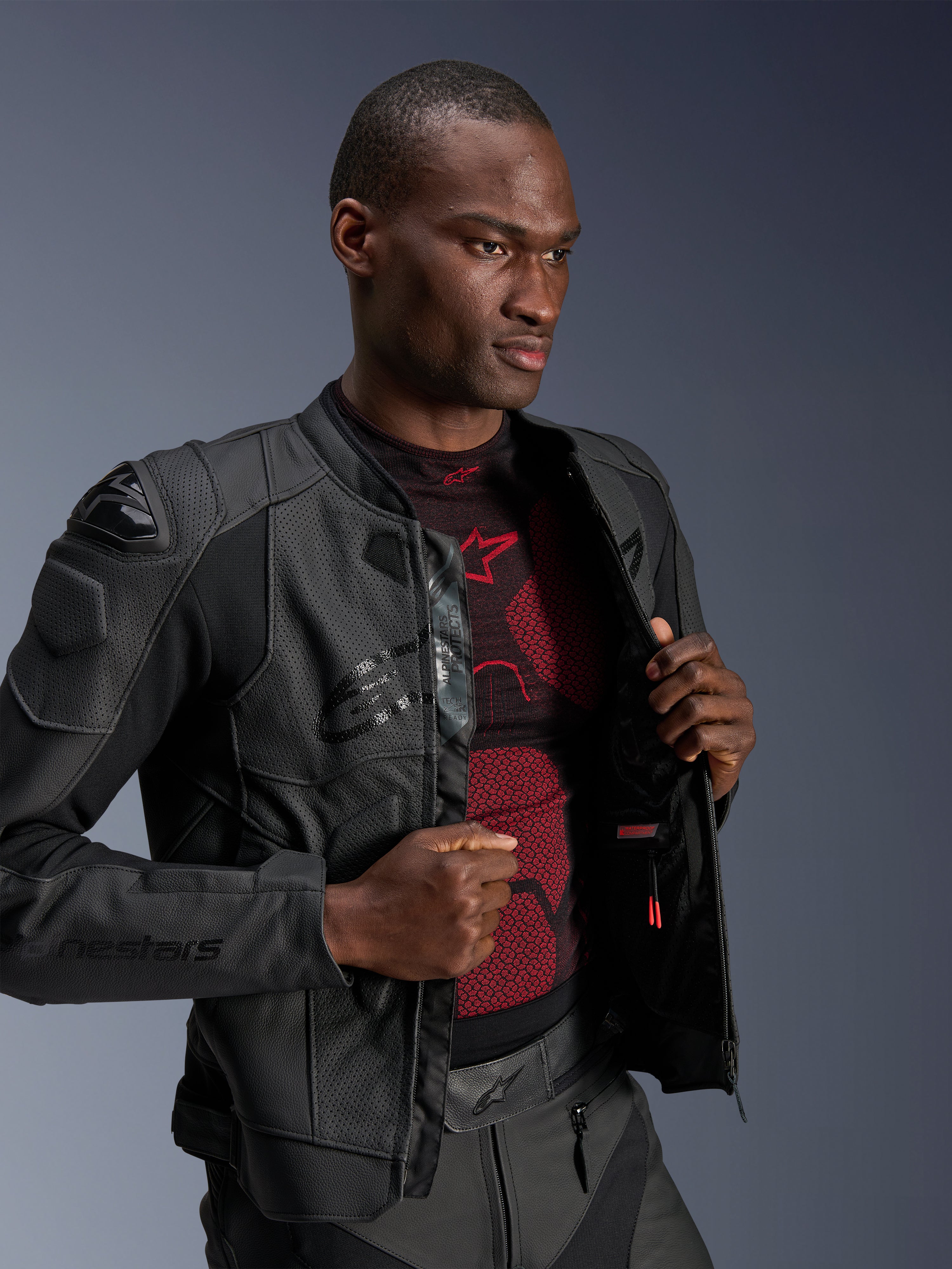 Alpinestars Dusk Airflow Lederjacke, Black Black, getragen von einem männlichen Modell über einer rot-schwarzen Basisschicht, mit perforierten Ledereinsätzen, externen DFS-Schulterschleifern und einer sportlichen Performance-Passform.