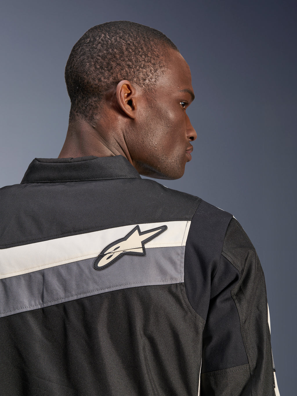Alpinestars T-Dyno WR Jacke, Textil-Motorradjacke, Schwarz, Dunkelgrau und Ecru, Nahaufnahme des oberen Rückens und der Schulter mit horizontalen Streifen und Alpinestars-Sternlogo, von einem Model getragen, Heritage-inspiriertes Design für Straßenfahrten