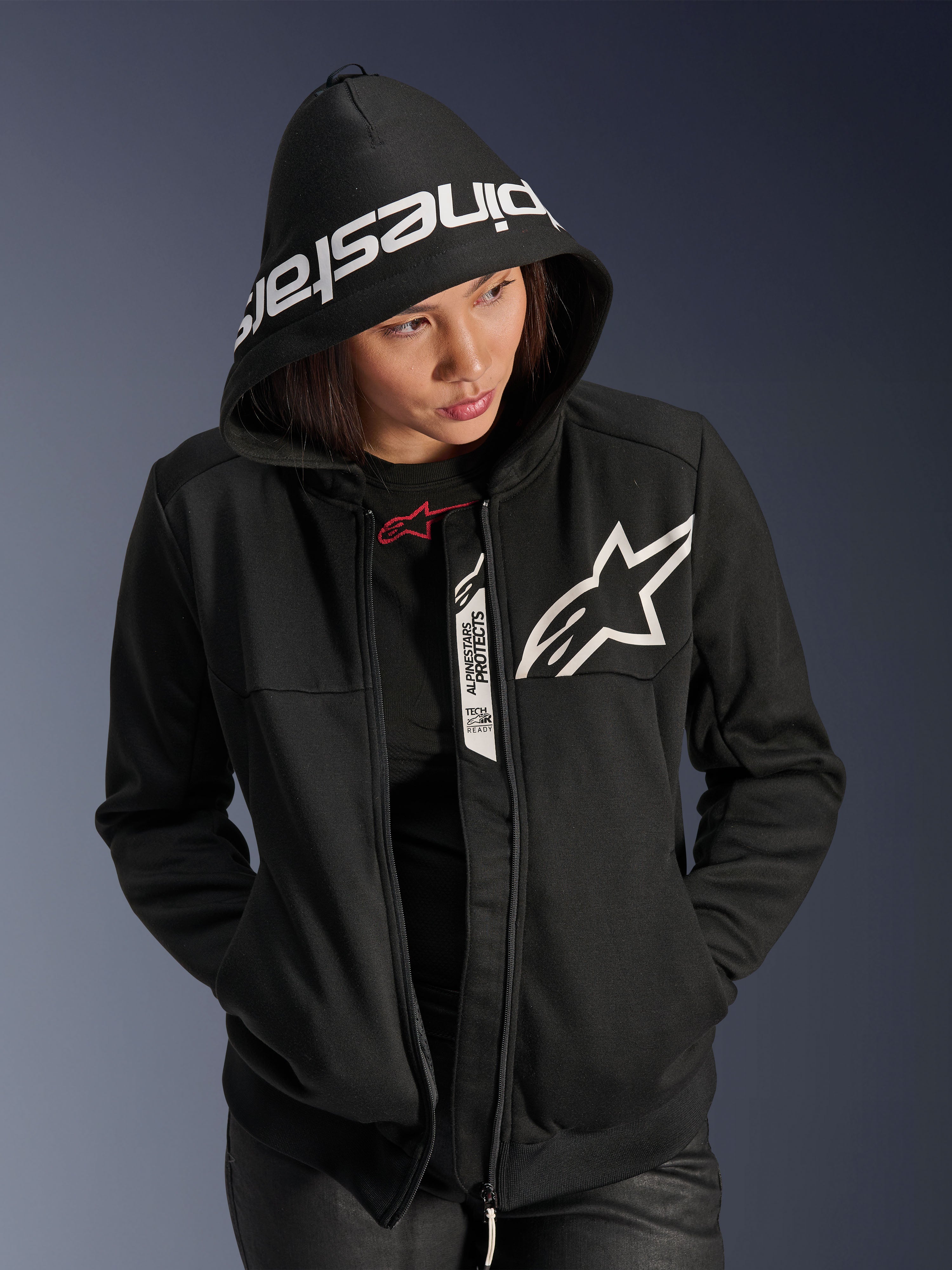 Alpinestars Stella Chrome V2 Sport Hoodie für Damen, Schwarz und Weiß, Motorrad-Kapuzenjacke mit weißem Stern-Logo auf der Brust und Alpinestars-Branding am Kapuzenrand, Frau mit aufgesetzter Kapuze, lässiger urbaner Sport-Style mit Tech-Air Ready-Details
