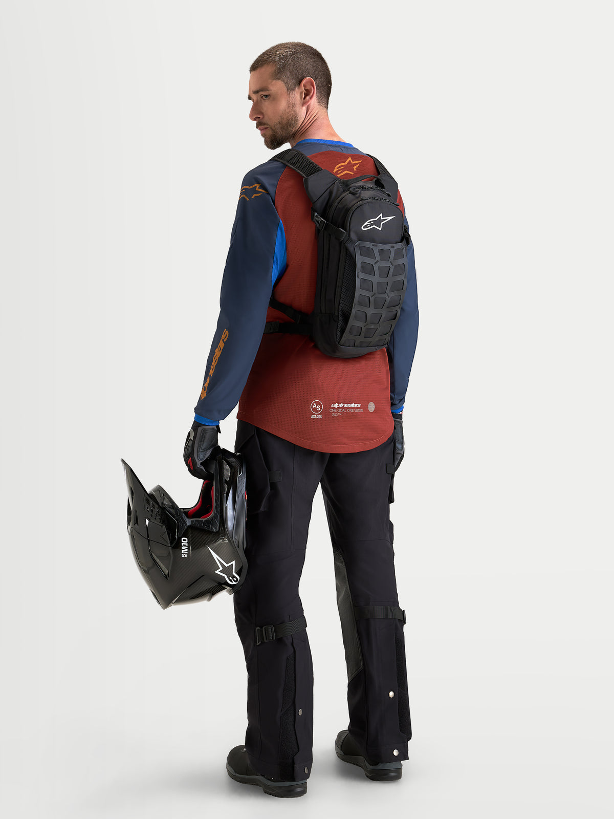 Alpinestars AT-9 Backpack, motocross backpack in black and deep gray, worn by a model from the rear view holding a helmet and wearing matching riding gear