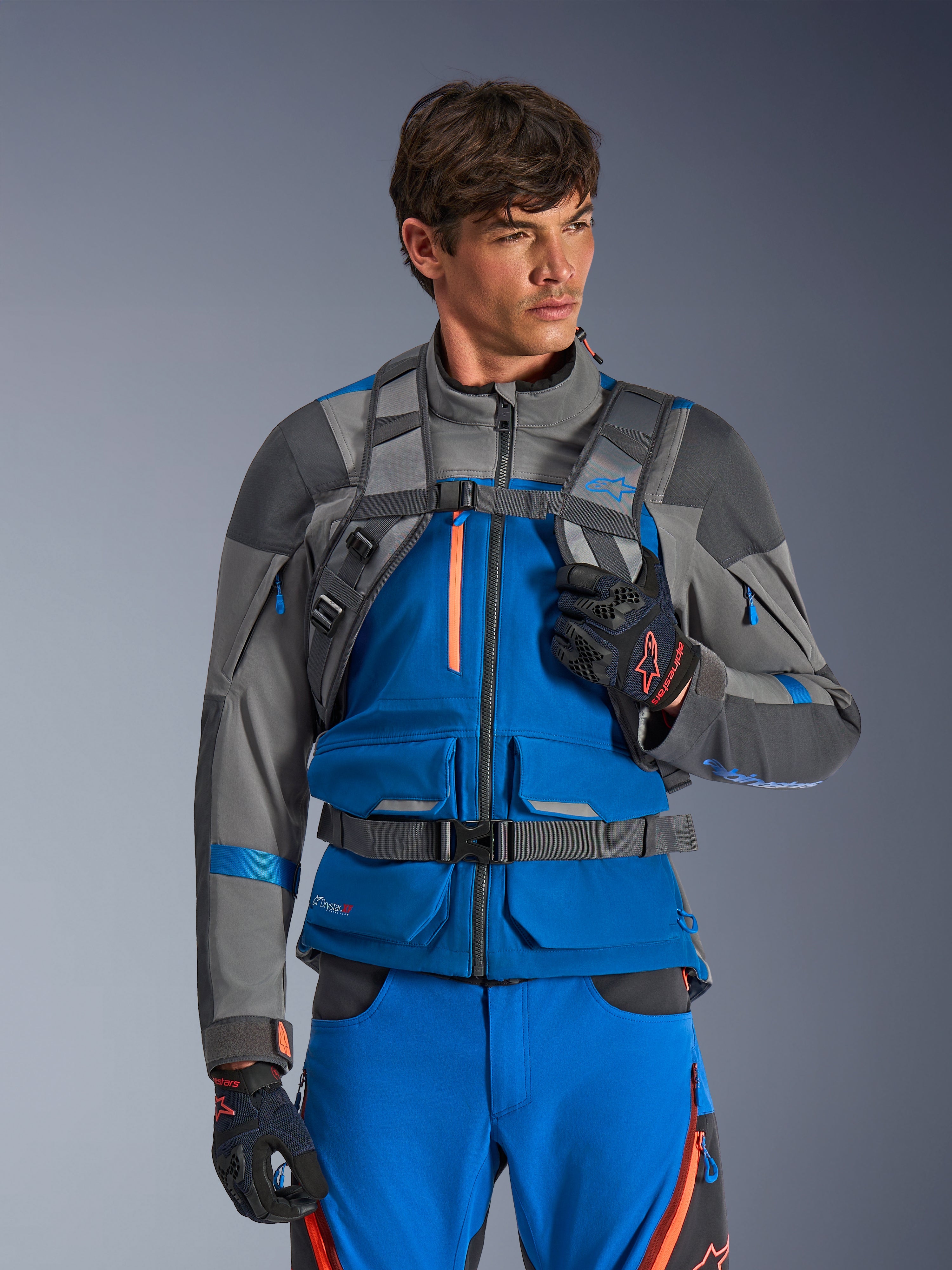 Alpinestars AT-15 Rucksack, Motorrad-Rucksack, Dunkelgrau, getragen von einem Fahrer in einer blau-grauen Adventure-Jacke und -Hose, mit technischen grauen Schultergurten, magnetischem Brustverschluss und Hüftgurt, robustes Design für Offroad-Fahrten