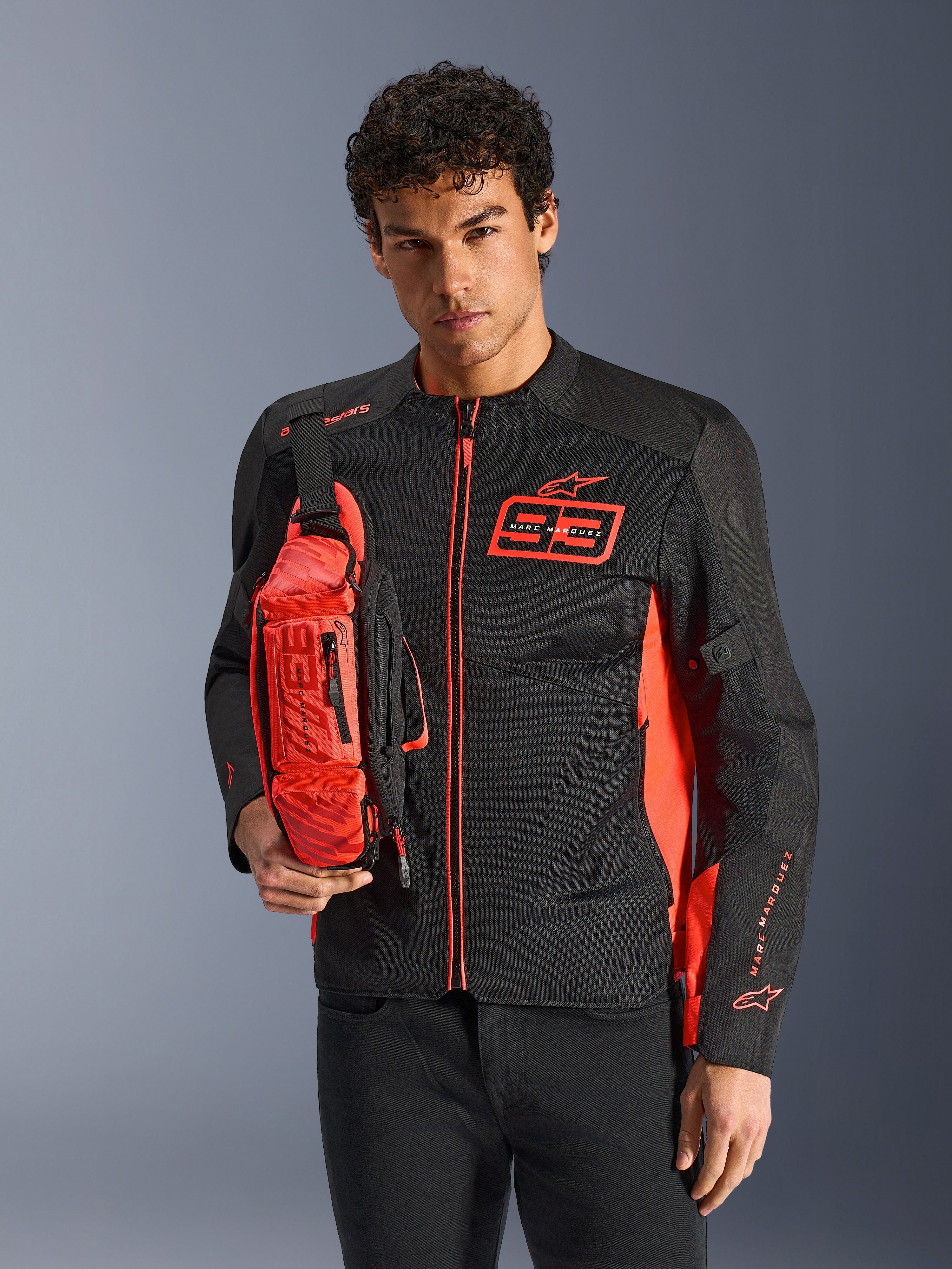Man wearing Alpinestars MM93 Waist Bag, Red Fluo and Black, styled as a crossbody bag over a matching black and red motorcycle jacket, featuring Marc Marquez 93 logo, sporty racing design